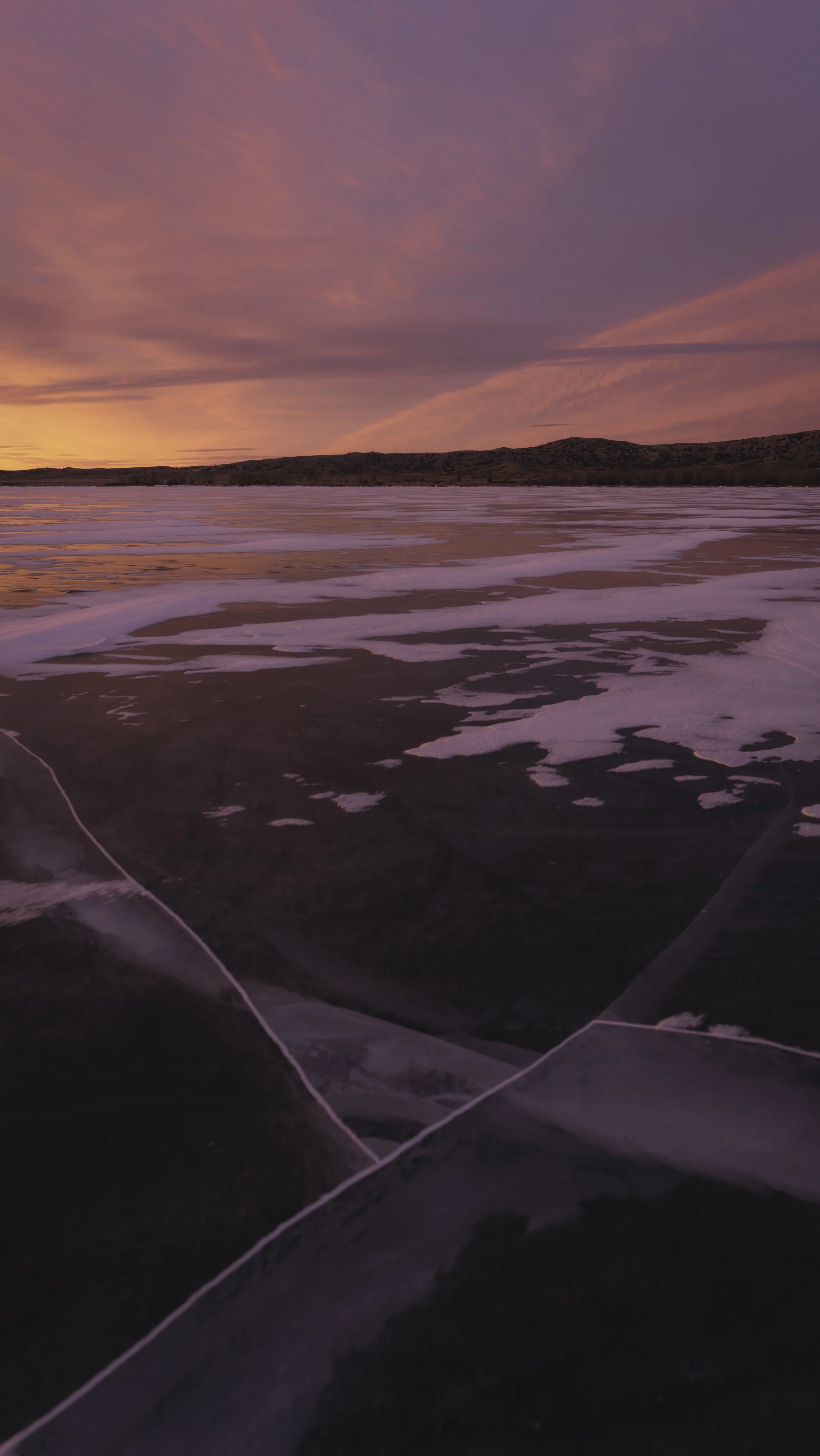 Frozen lake at sunset with pink, purple, orange hues in the sky and ice with cracks in the foreground.