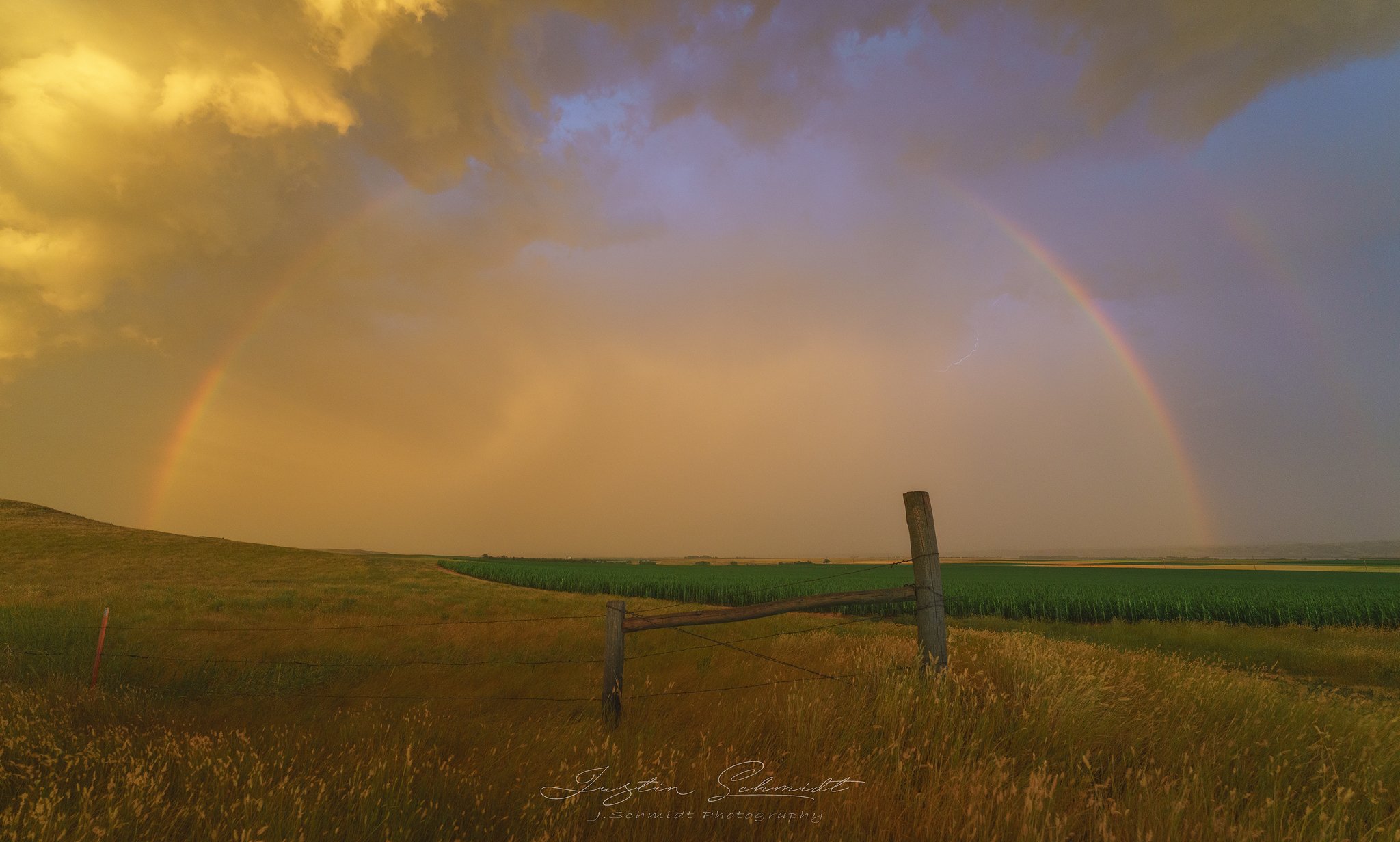 A double rainbow across a grassy field, with a fence in the foreground and agricultural land in the distance, under a partly cloudy sky with a lightning bolt.