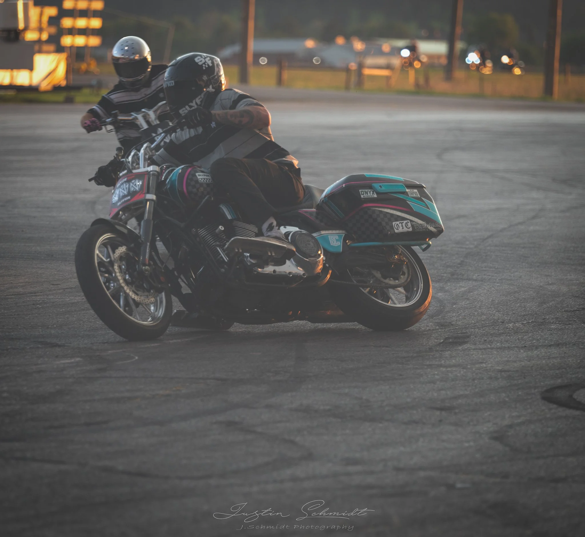 Two motorcyclists riding on a curved asphalt road at dusk, wearing helmets and riding gear, with blurred background of cars and streetlights.