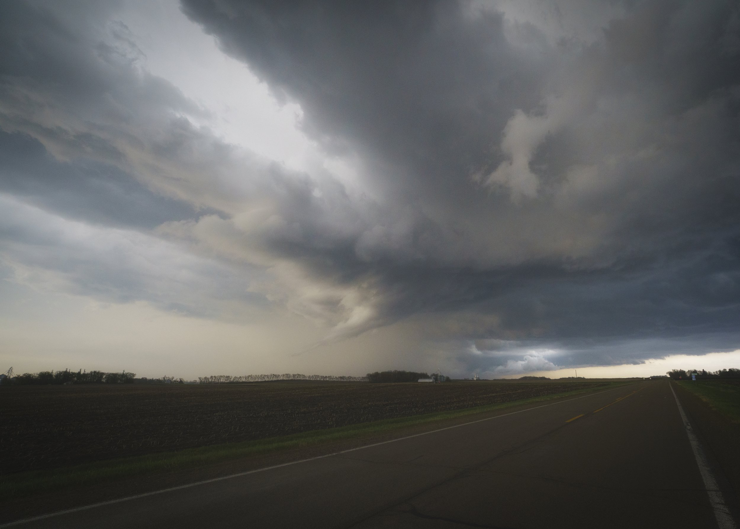 Overcast sky with dark storm clouds over an empty rural road and field, indicating an approaching storm.