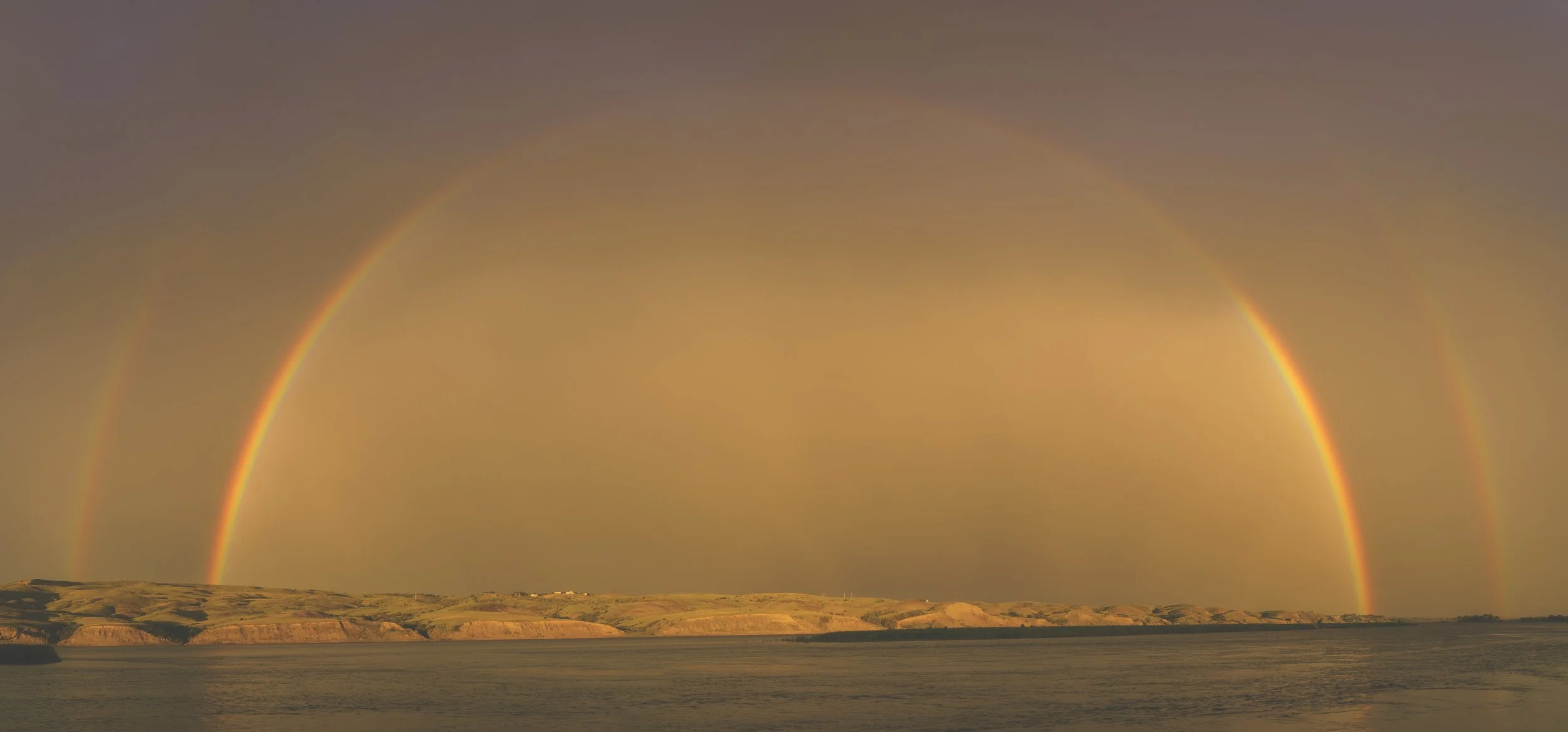 A double rainbow arching over a coastal landscape at sunset, with water in the foreground and hills in the background.