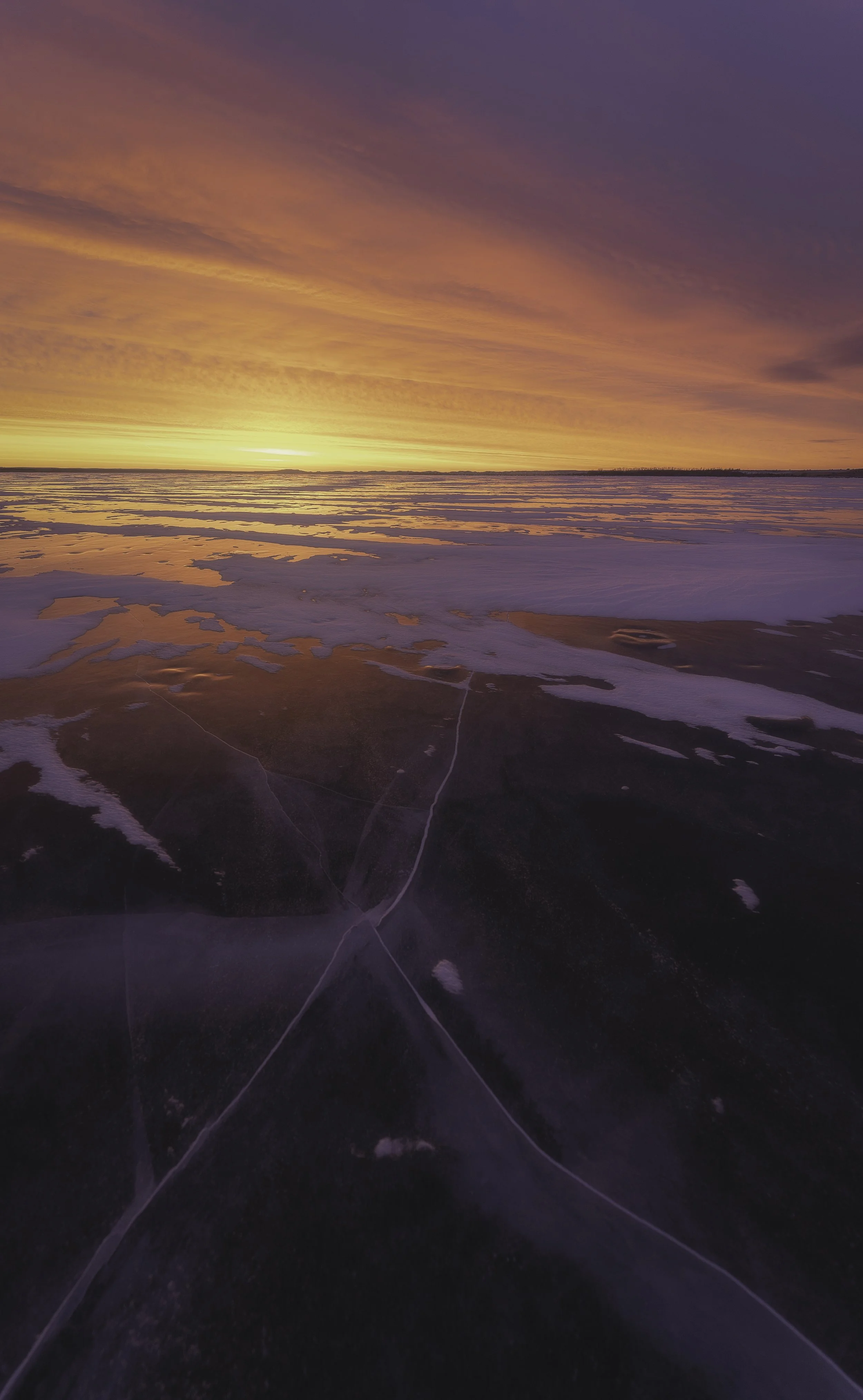 A sunset over a large frozen lake with cracked ice and patches of snow, showing a colorful sky with orange, pink, and purple hues.