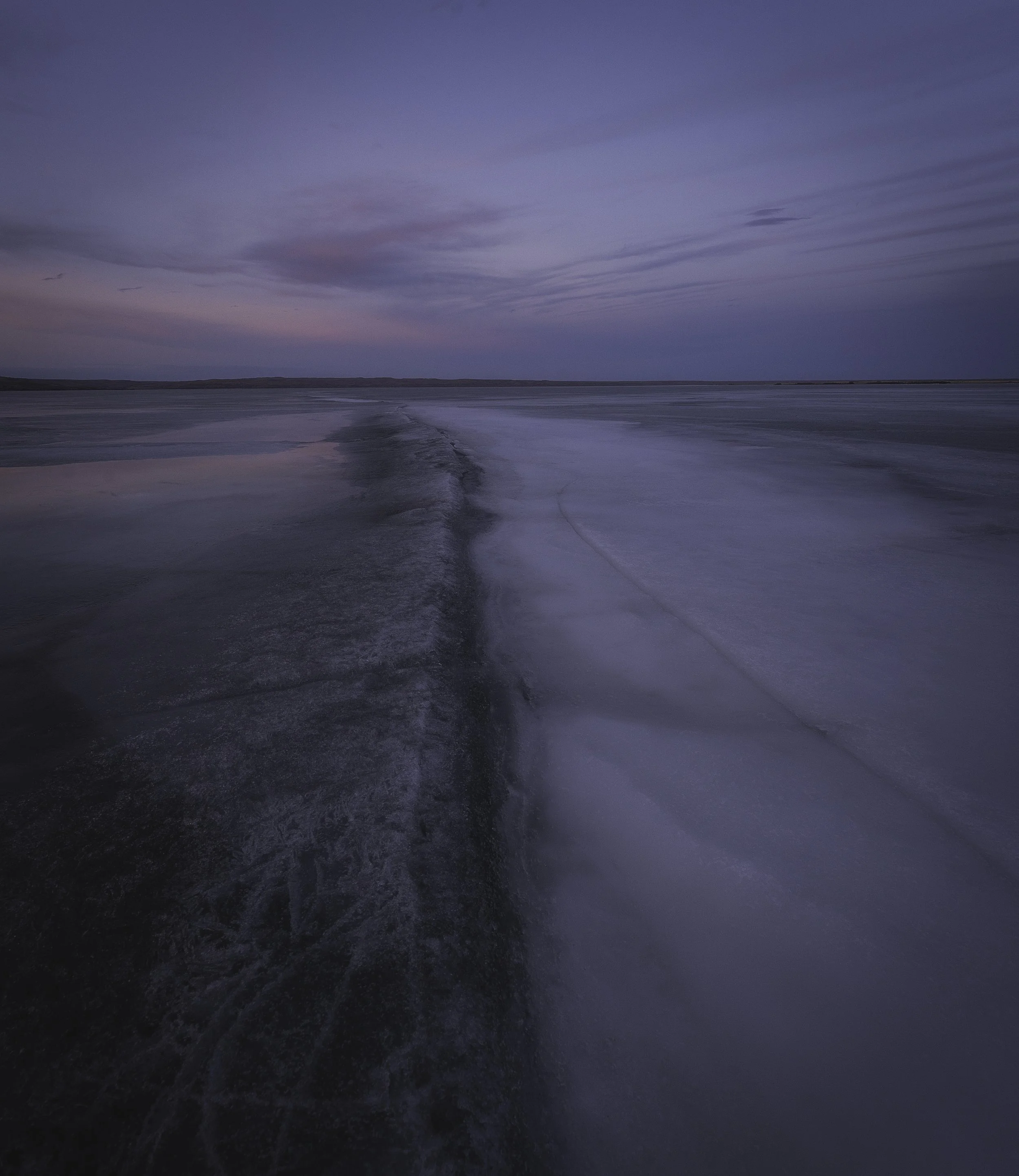 Frozen lake with a trail of water and icy surface under a pink and purple sky at dusk.