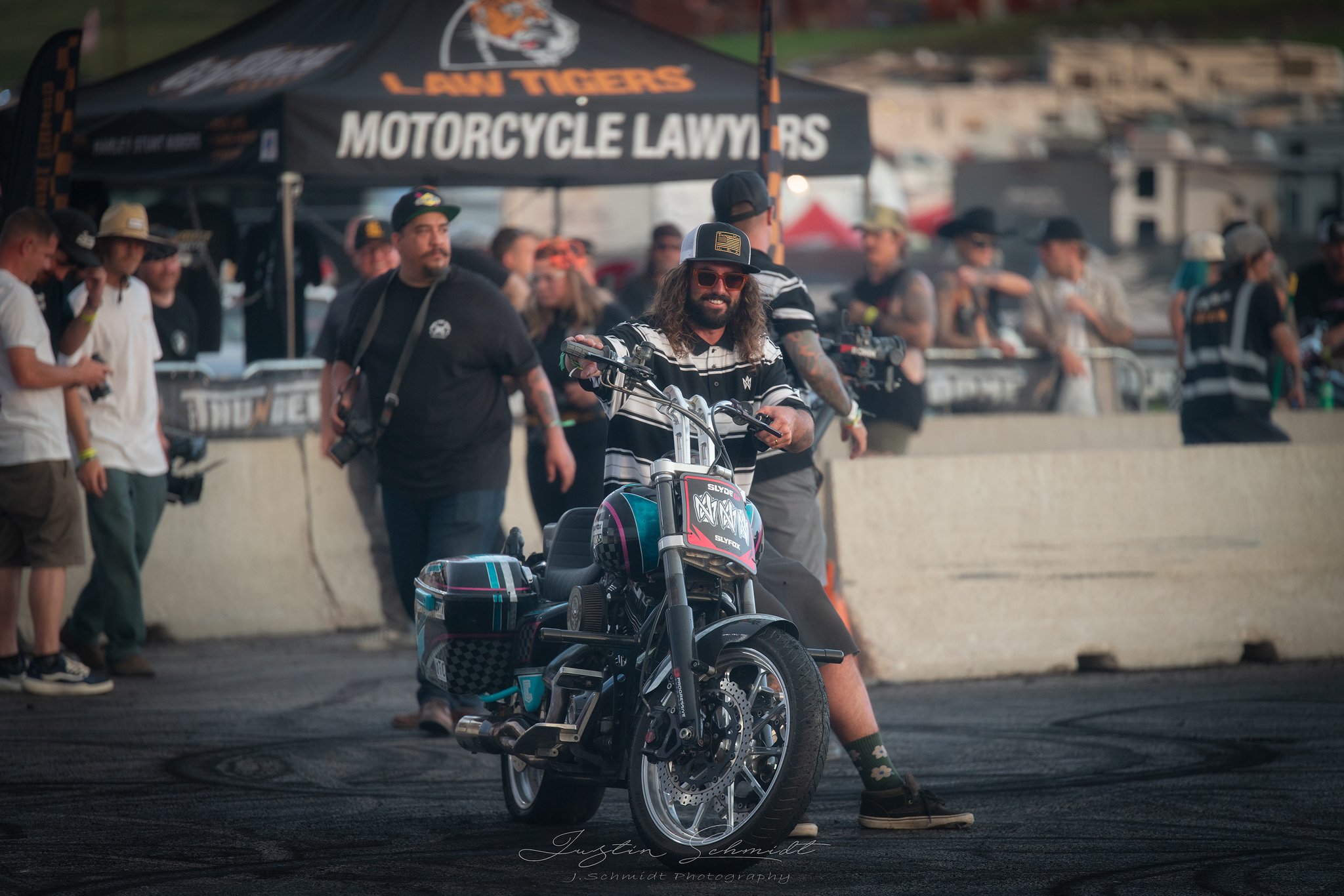 A man with curly hair, a beard, sunglasses, and a hat is sitting on a black motorcycle at a motorsport event. There are other people, some with cameras, in the background. Tents and banners related to motorcycle racing are visible.