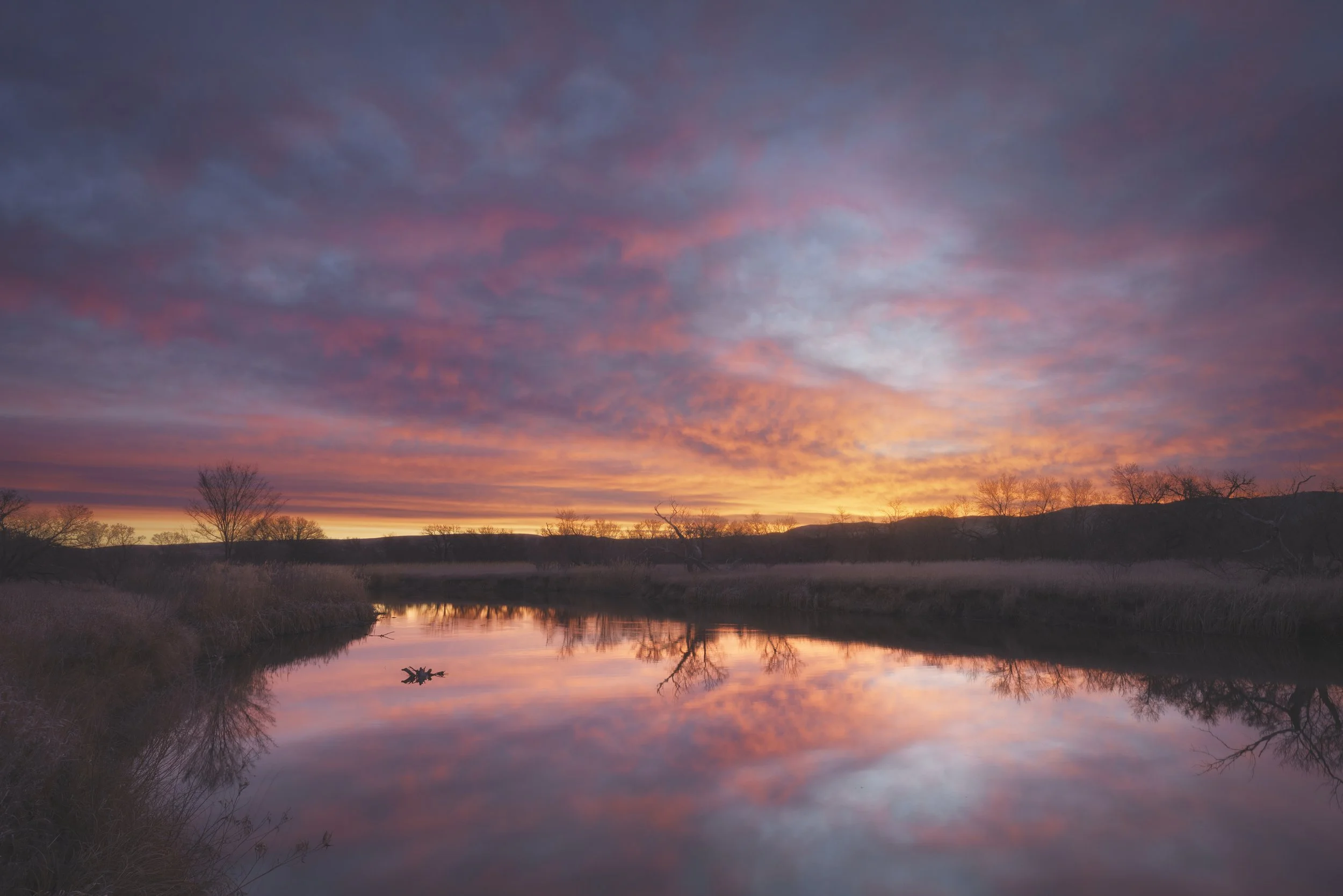 A serene sunset over a calm river, with a colorful sky of pink, purple, and orange clouds, and silhouettes of trees along the riverbank.