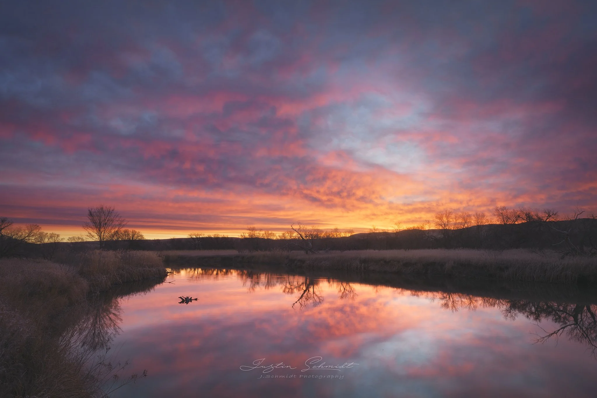 Sunset over a river with colorful clouds in the sky and bare trees reflected in the water.
