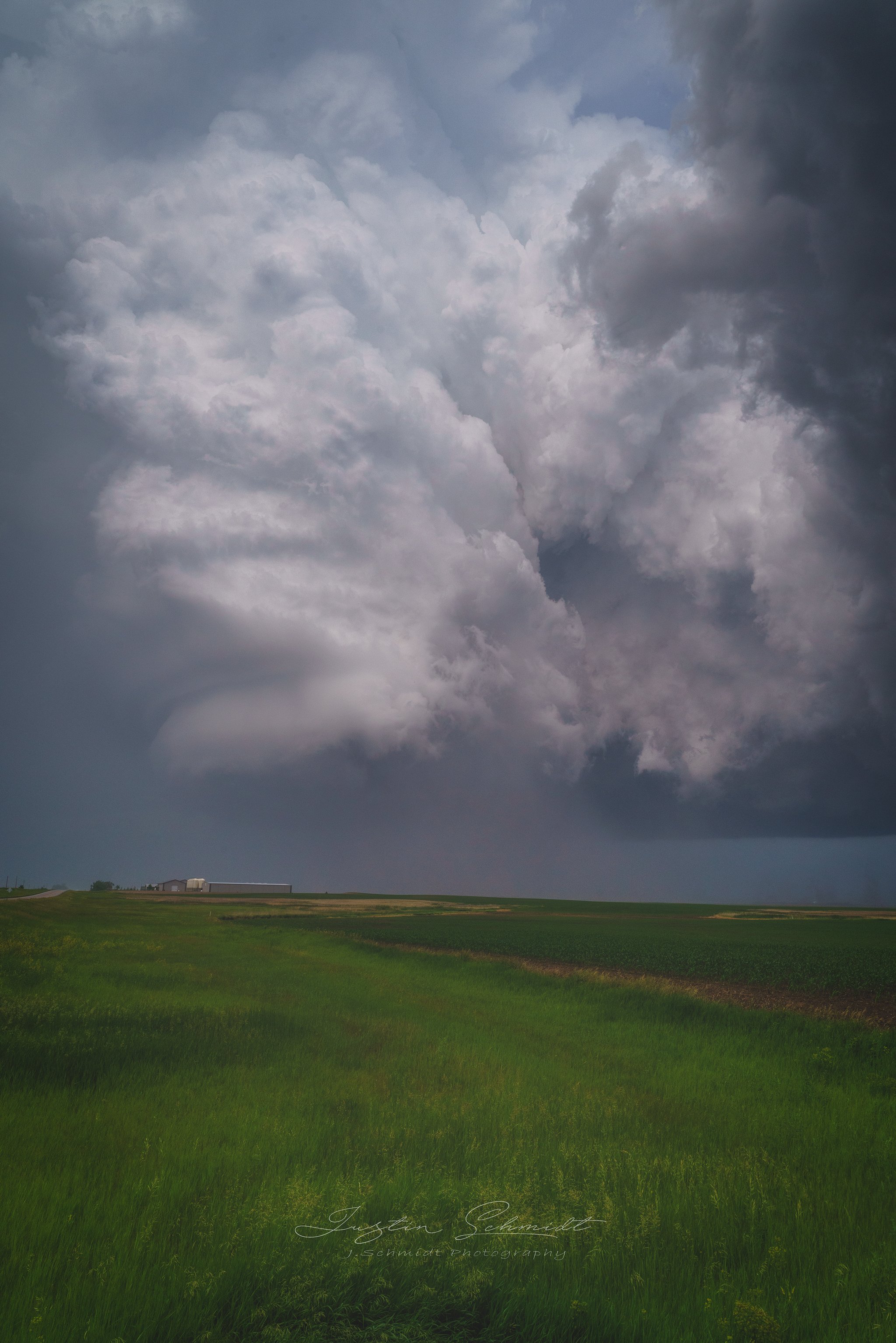 A large, dark storm cloud over a green field with a farm building in the distance.