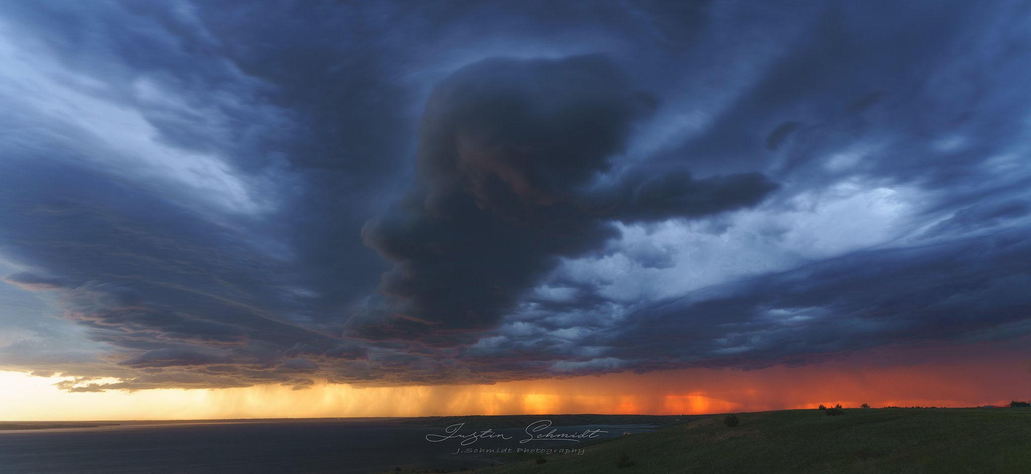 Dark storm clouds over a flat landscape during sunset, with rain visible at the horizon.