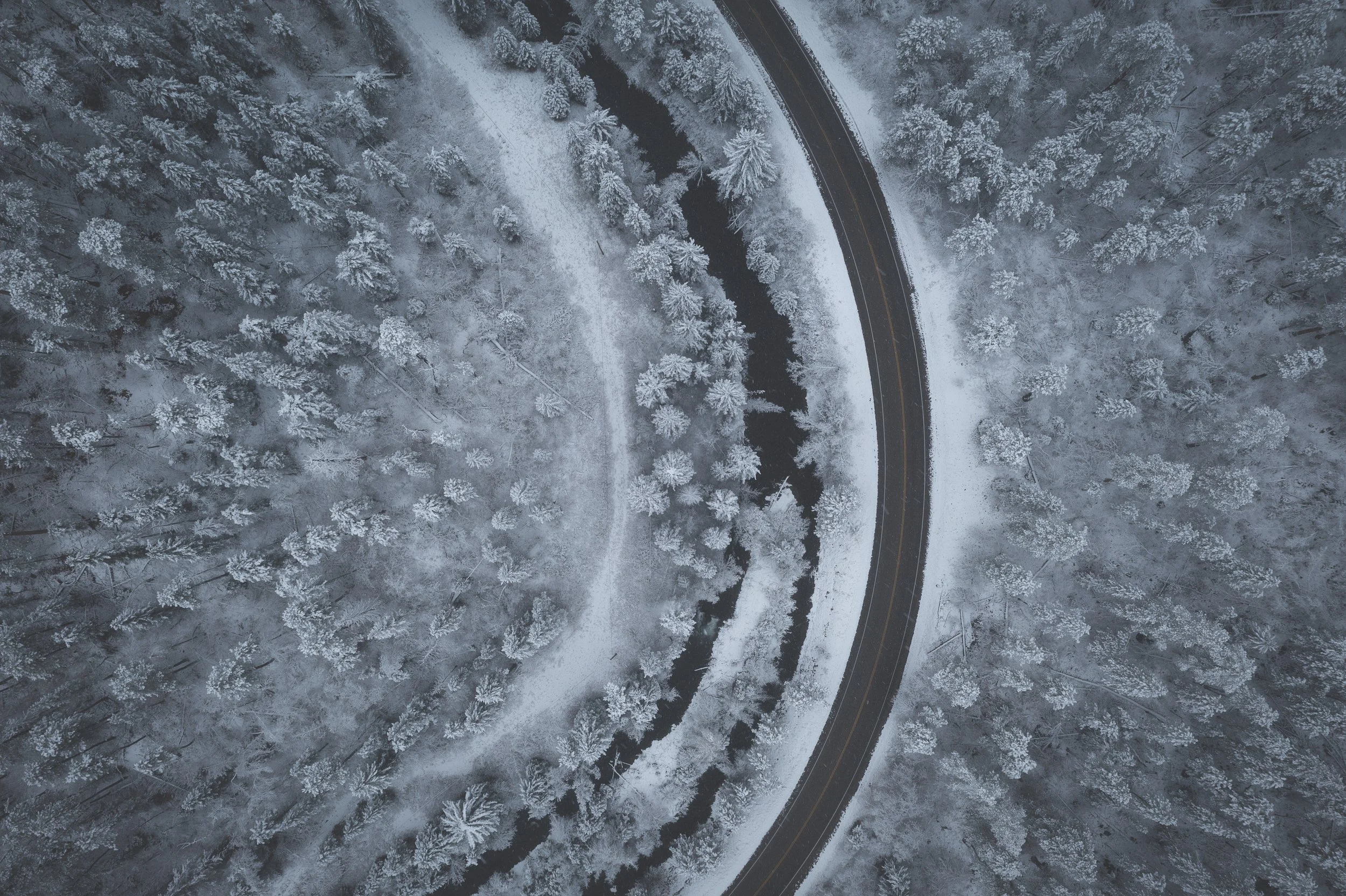 Aerial view of a winding road cutting through a snow-covered forest with trees laden with snow.