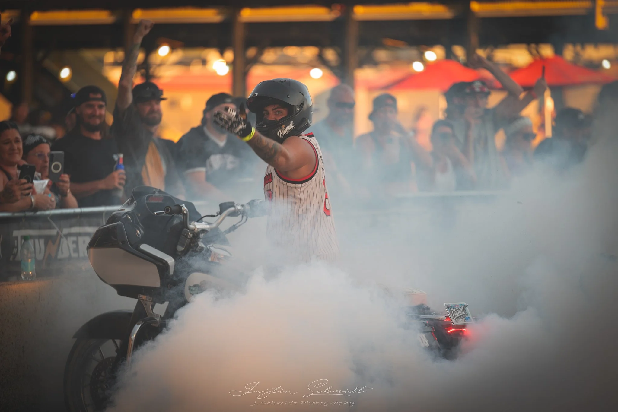 A person wearing a helmet and racing jersey standing next to a motorcycle, surrounded by smoke at a motorsport event with a crowd of spectators in the background.