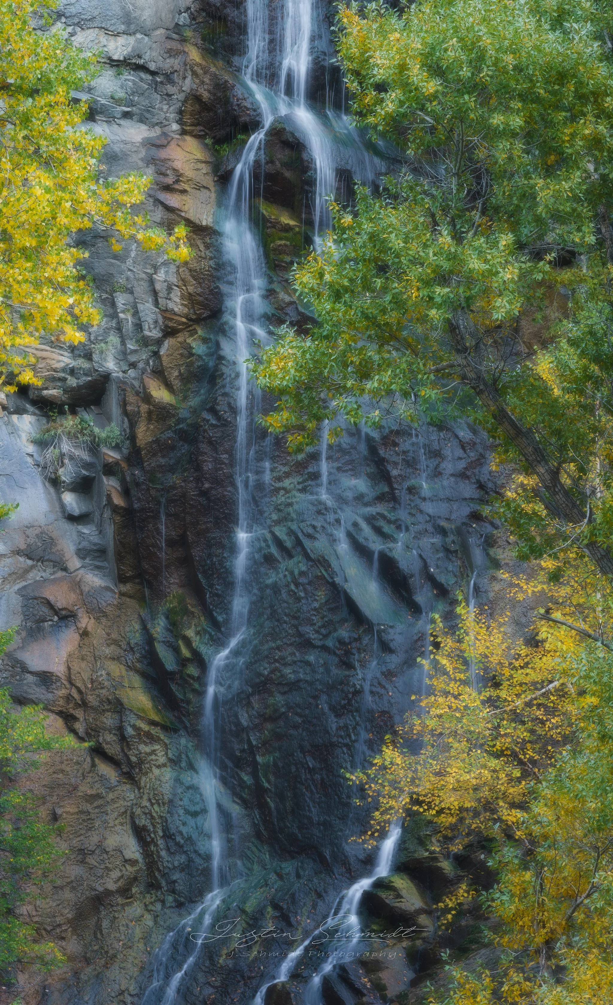 A waterfall cascading down a rocky cliff surrounded by green and yellow autumn foliage.