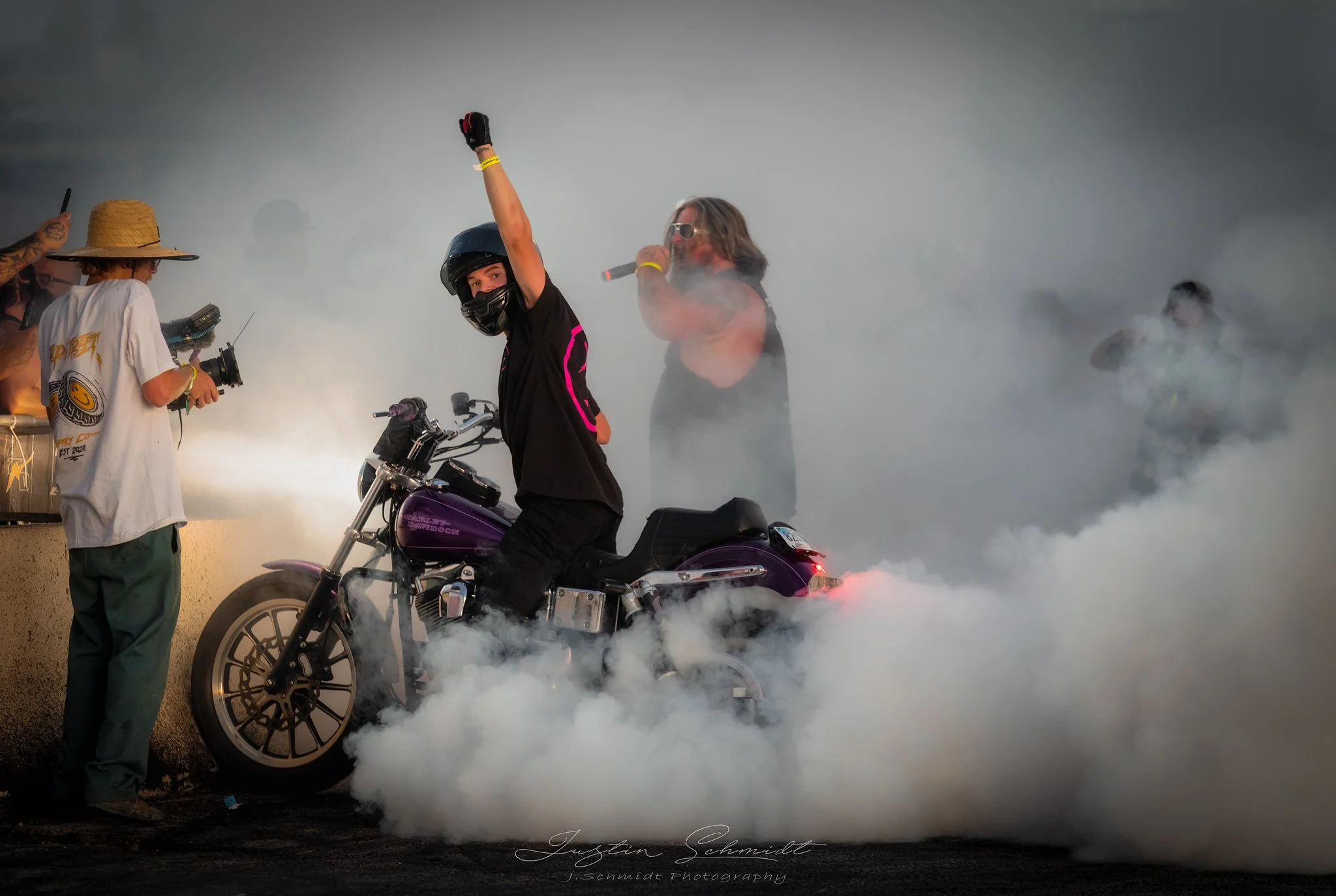 A motorcyclist wearing a helmet and black outfit raising one arm, sitting on a purple motorcycle, surrounded by white smoke. A woman behind the motorcyclist holds a microphone, and there are spectators, some with cameras, in the background.
