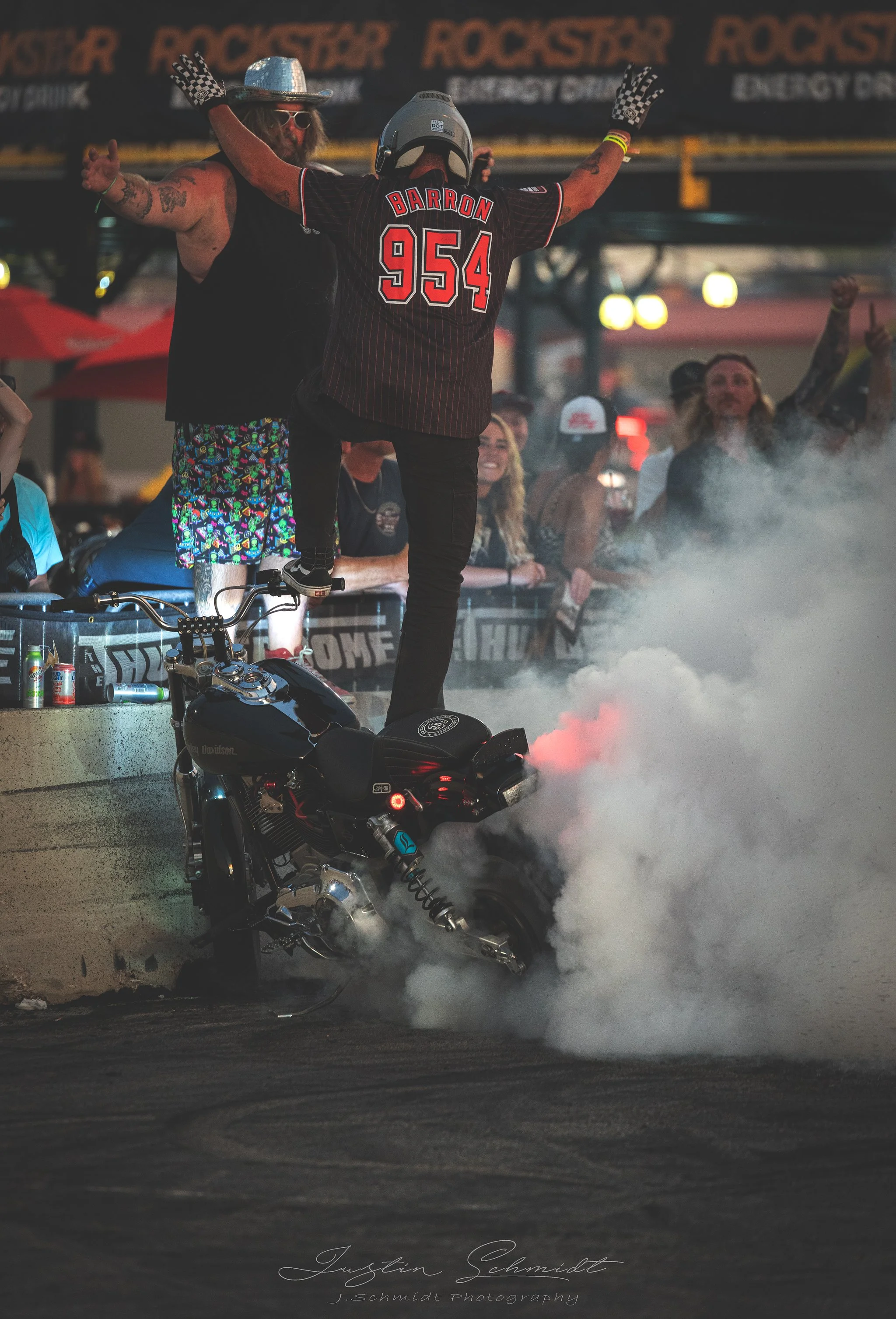 A man on a motorcycle is doing a burnout at a racing event, with smoke billowing from the rear tire and a crowd of spectators watching behind a barrier.