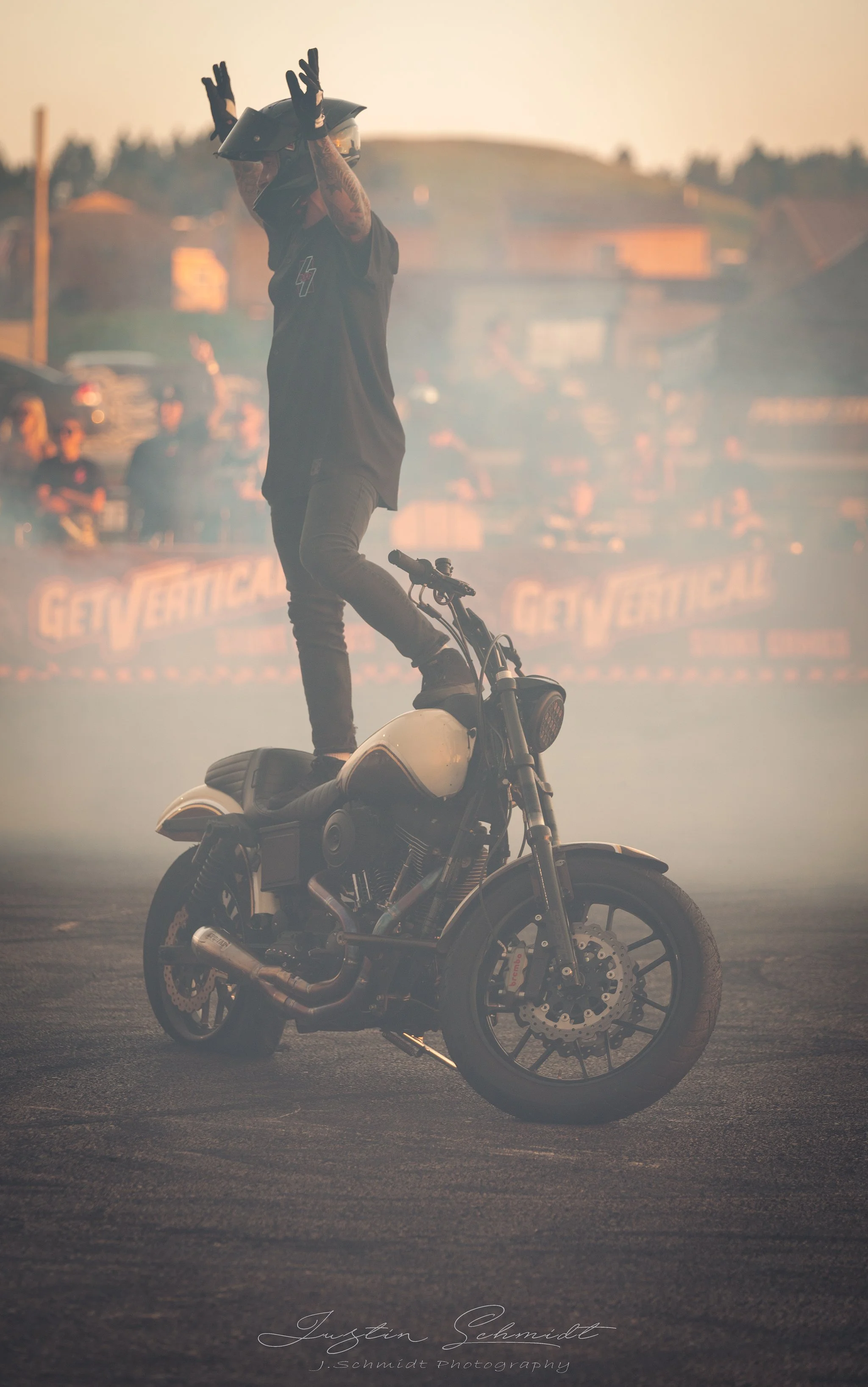 A person wearing motorcycle gear and a helmet is balancing on a motorcycle while standing on the seat with arms raised. The scene is smoky with a background of cars and people, possibly at a motorsport event or stunt show.
