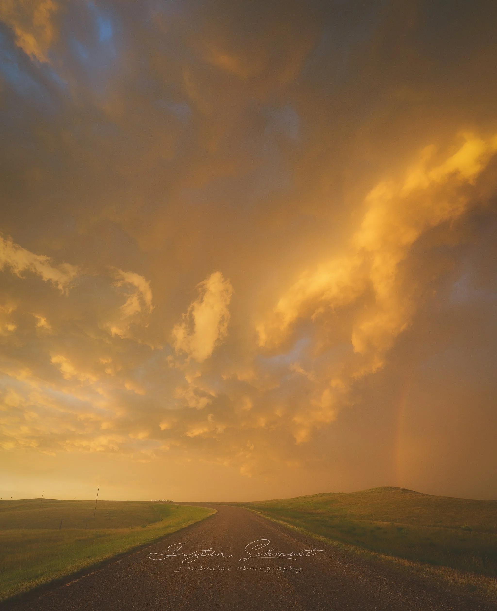 A rural road stretching into the distance under a dramatic sky during sunset, with orange and yellow clouds.