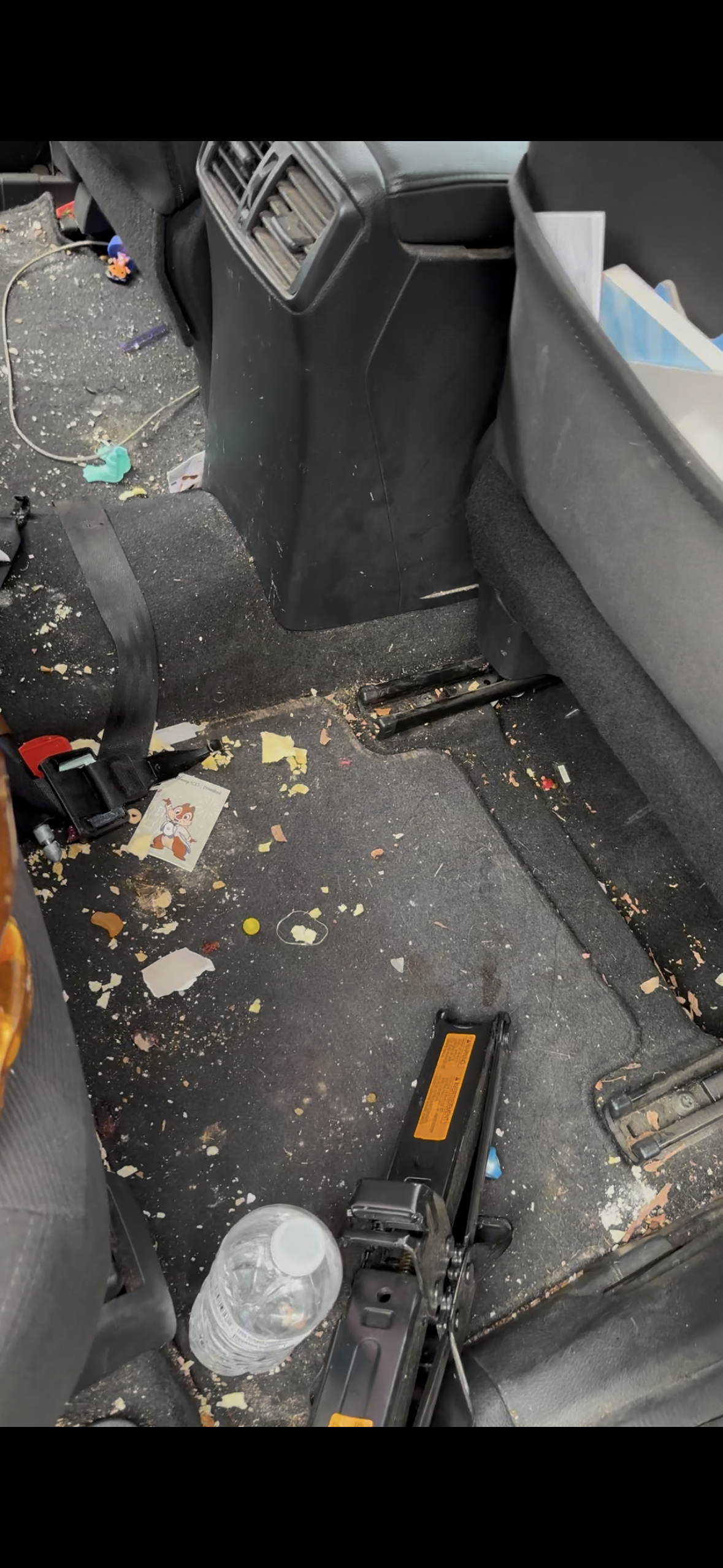Inside a messy car with spilled snacks and debris on the floor, a plastic water bottle laying on the carpet. Disorganized back seats with papers and toys visible.