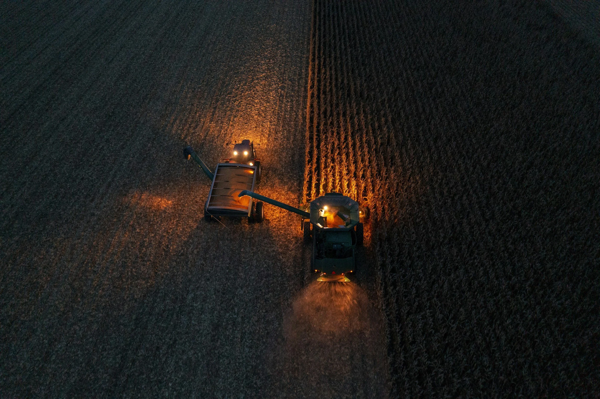 An aerial view of a tractor working in a large field at night, with lights illuminating the crop rows.
