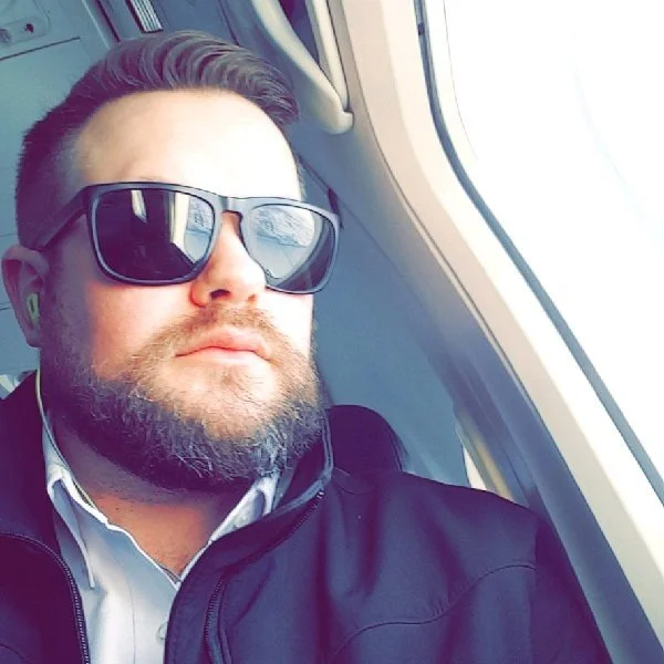 A man with a beard wearing sunglasses, a white shirt, and a black jacket, sitting by an airplane window.