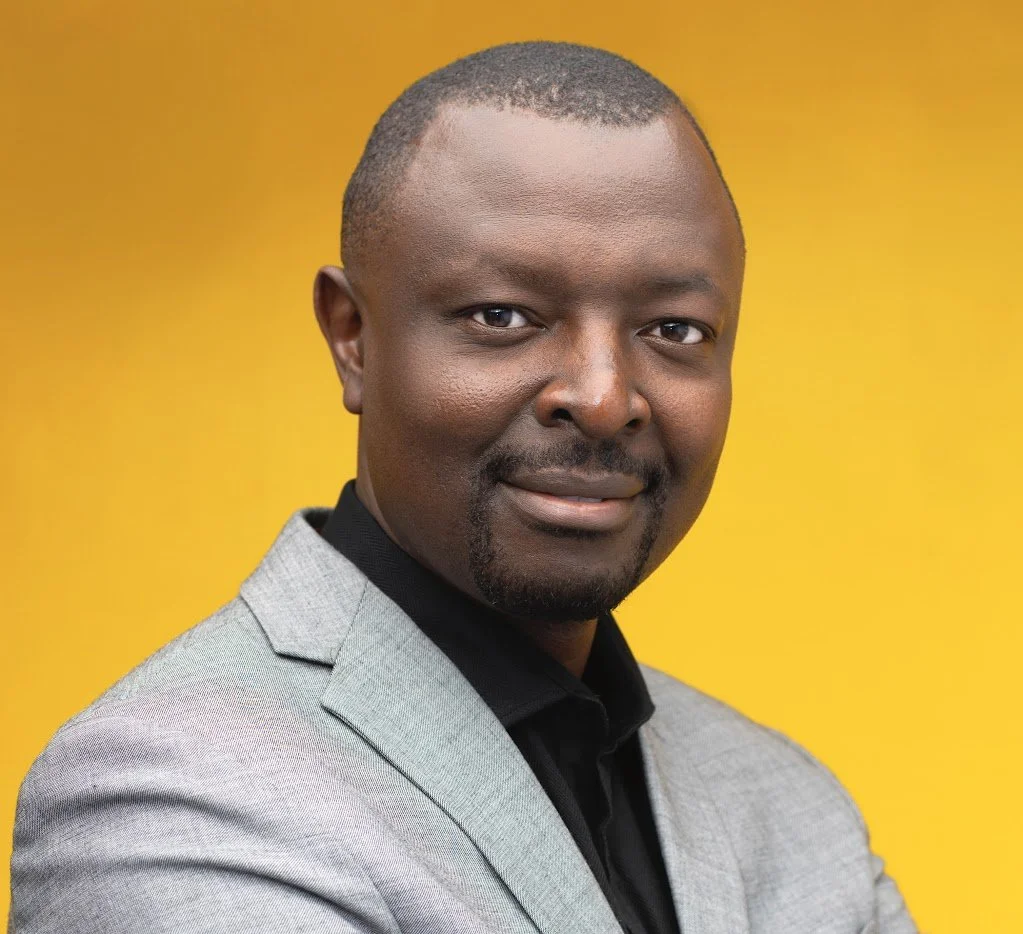 A portrait of a Black man in a gray blazer and black shirt against a yellow background.