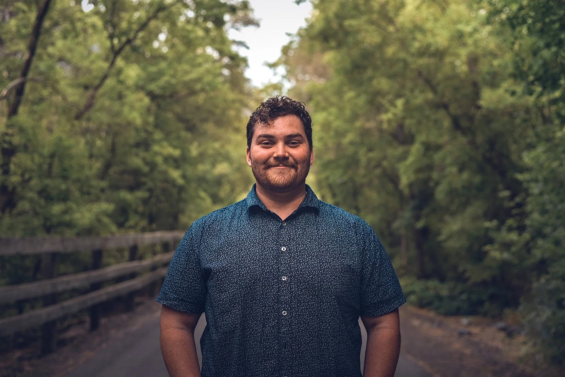 A man with dark curly hair and a beard, dressed in a short-sleeved button-up shirt, stands on a wooded forest path with trees and greenery in the background.