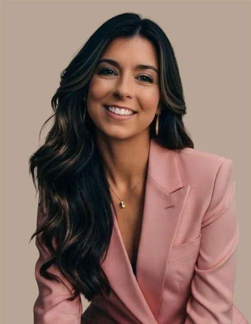 A woman with long, dark, wavy hair smiling, wearing a pink blazer and gold jewelry, against a neutral background.