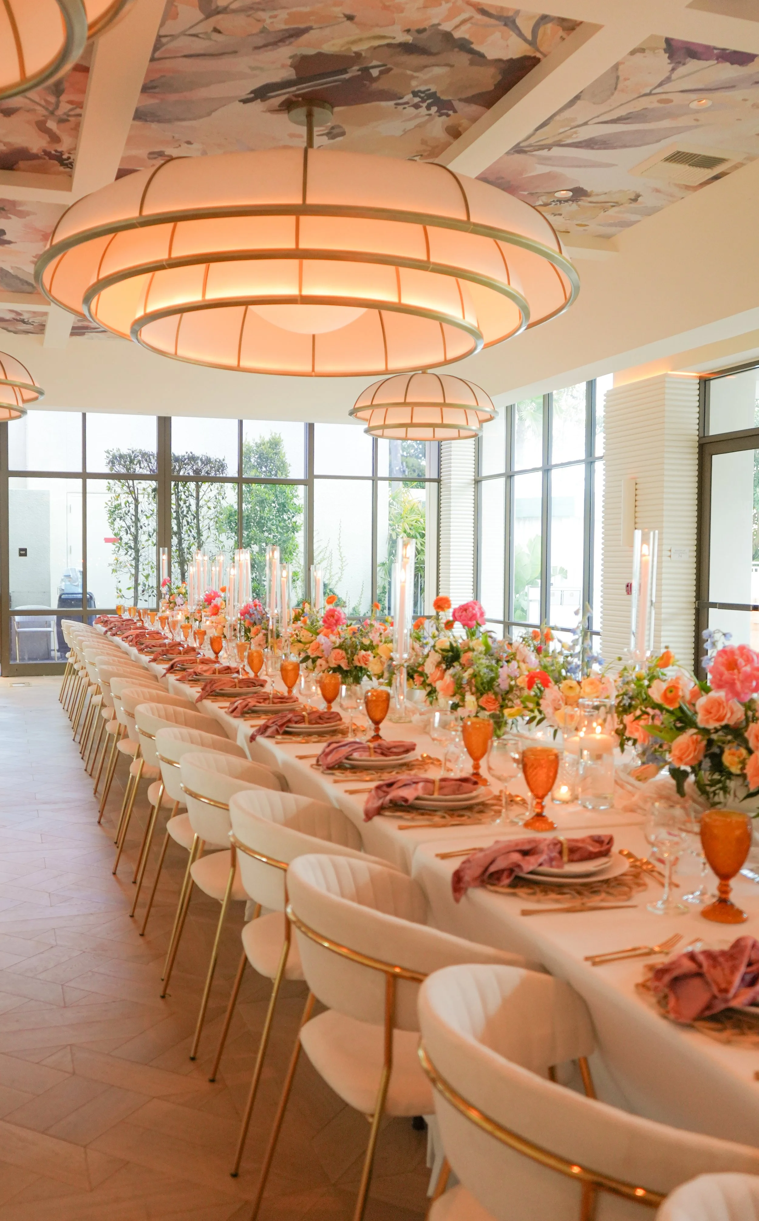 Long dining table decorated with pink and peach flowers, candles, and glasses in a bright, modern room with large windows and multiple round pendant lights.