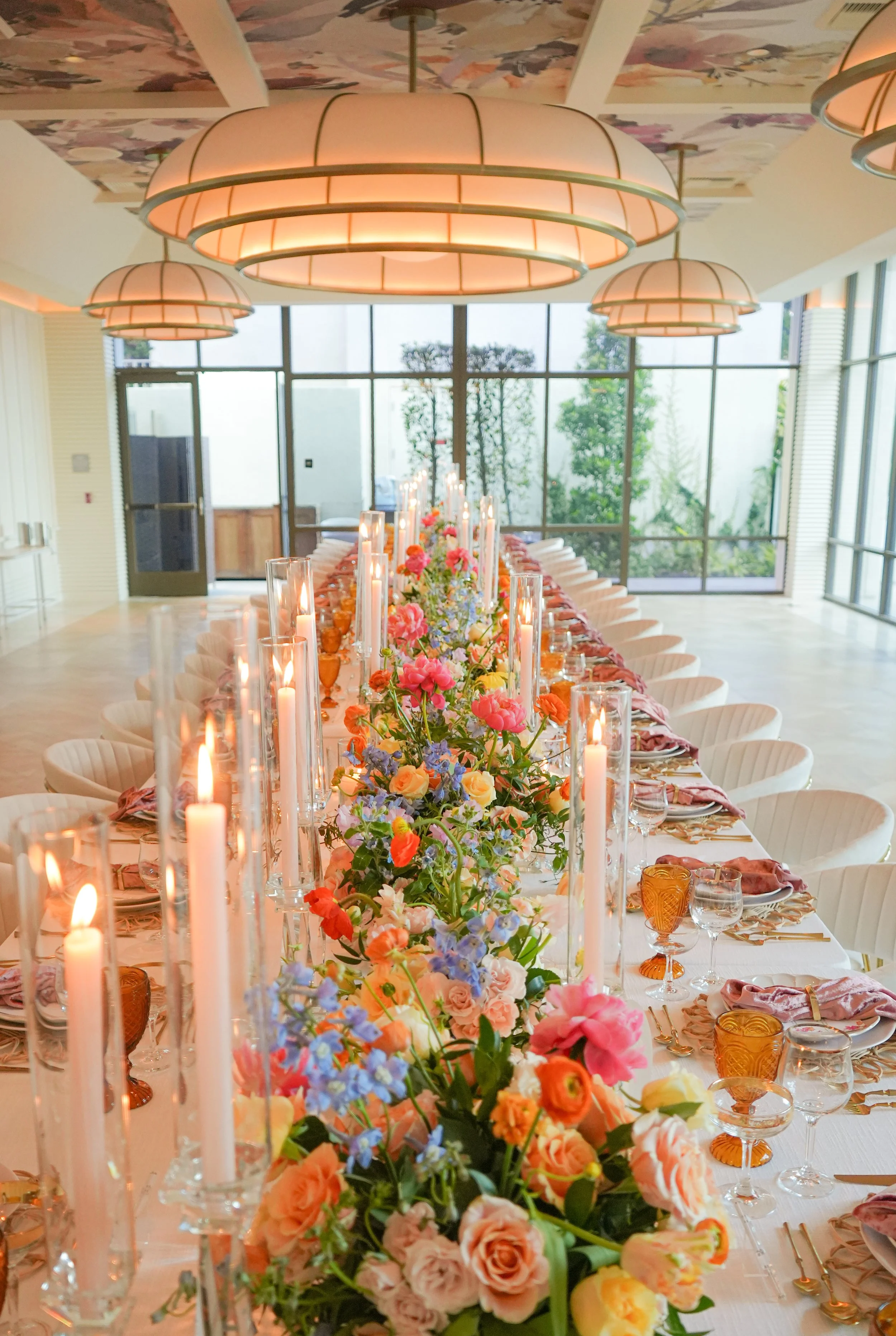Long dining table decorated with colorful flowers, candles in glass holders, and pink napkins in a bright, modern venue with large windows and round pendant lights.