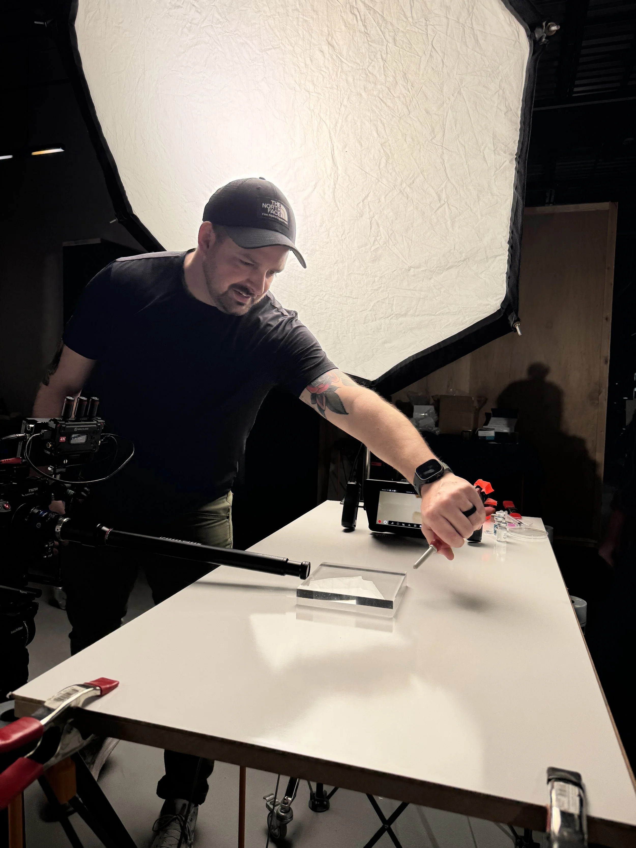 A man operating filming equipment in a studio, with a large softbox light overhead and a white table with various tools and equipment.