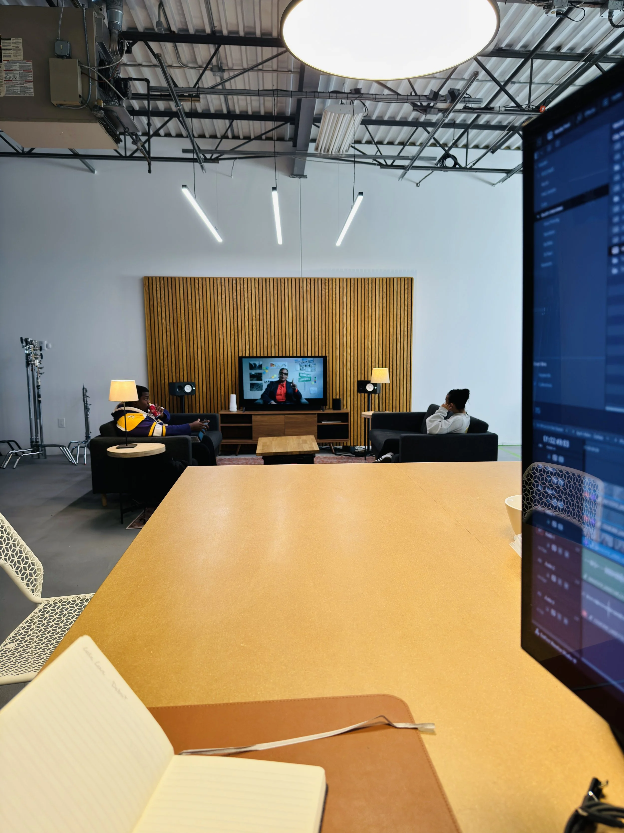 View of a modern office or studio with a large orange table in the foreground, an open notebook, and a computer screen. In the background, three people are seated on black couches watching television mounted on a wooden panel wall, with lighting and studio equipment visible above.