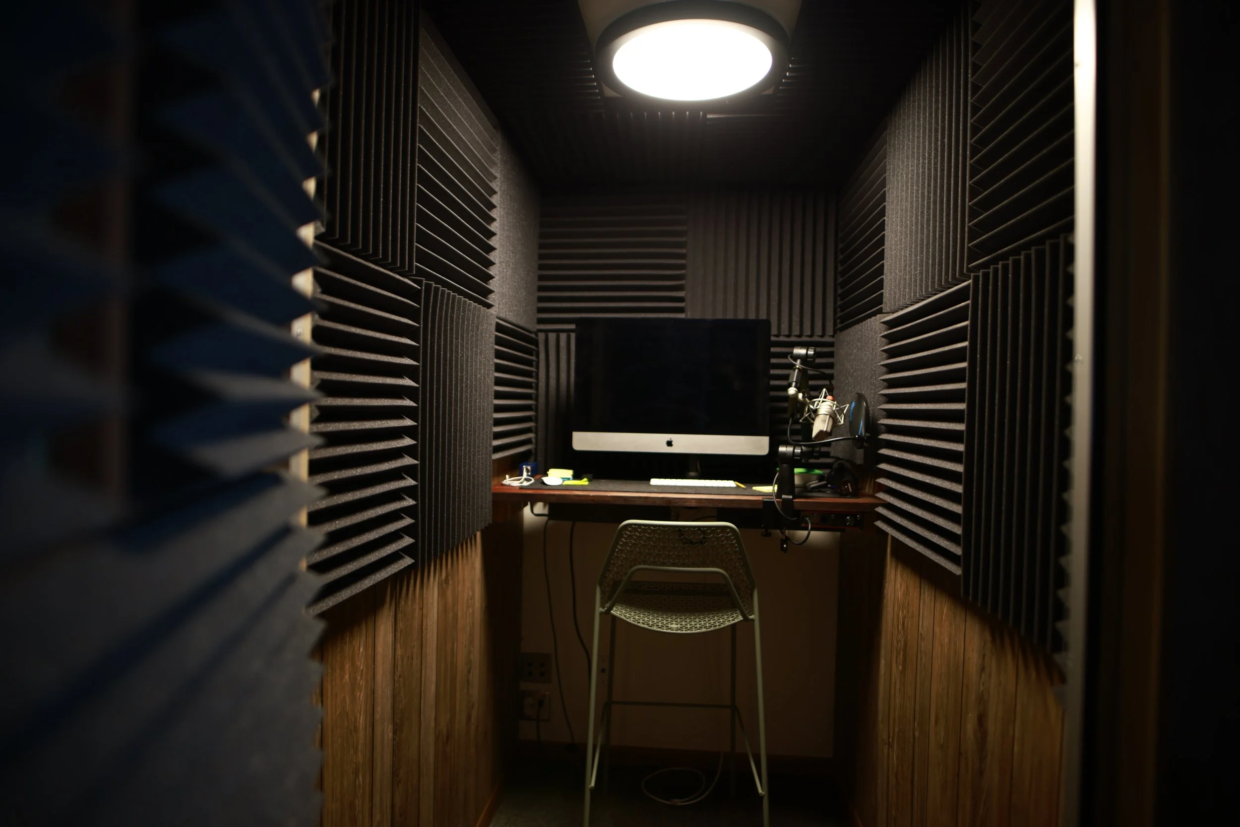 Recording or soundproofing booth with black acoustic foam walls, a computer monitor on a desk, a chair, and audio recording equipment.