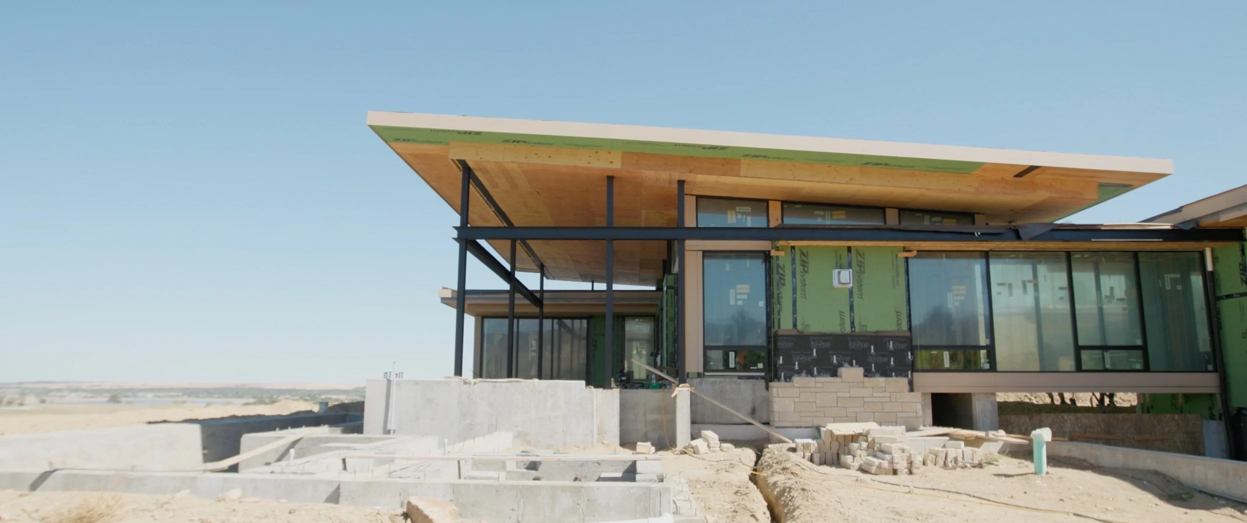 Modern house under construction with large glass windows, wooden roof, and metal framework, in a sparsely developed area under a clear blue sky.