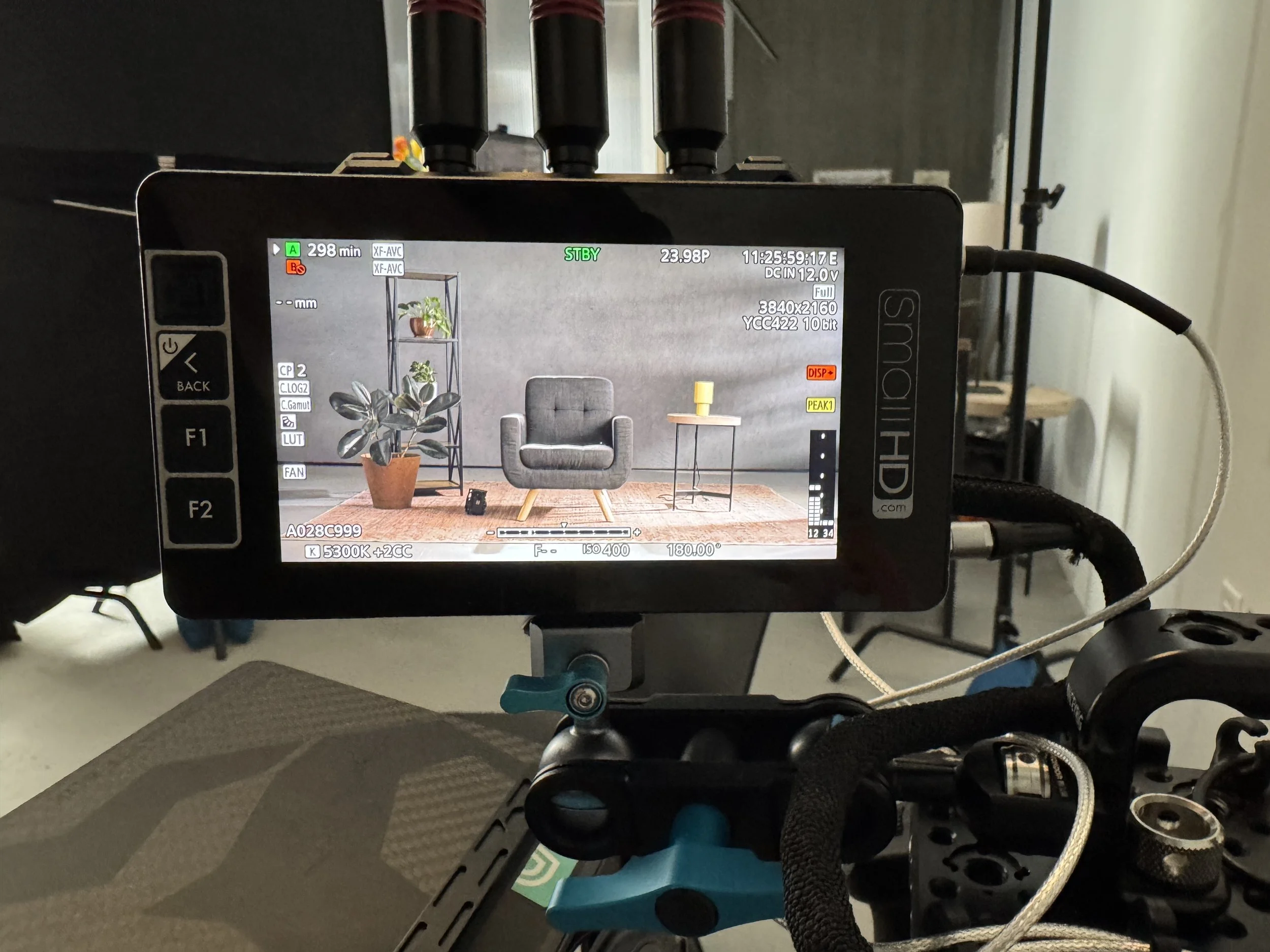 Camera monitor displaying a scene with a gray armchair, a small side table with a yellow object on it, a potted plant, and a metal shelf with plants against a gray wall.