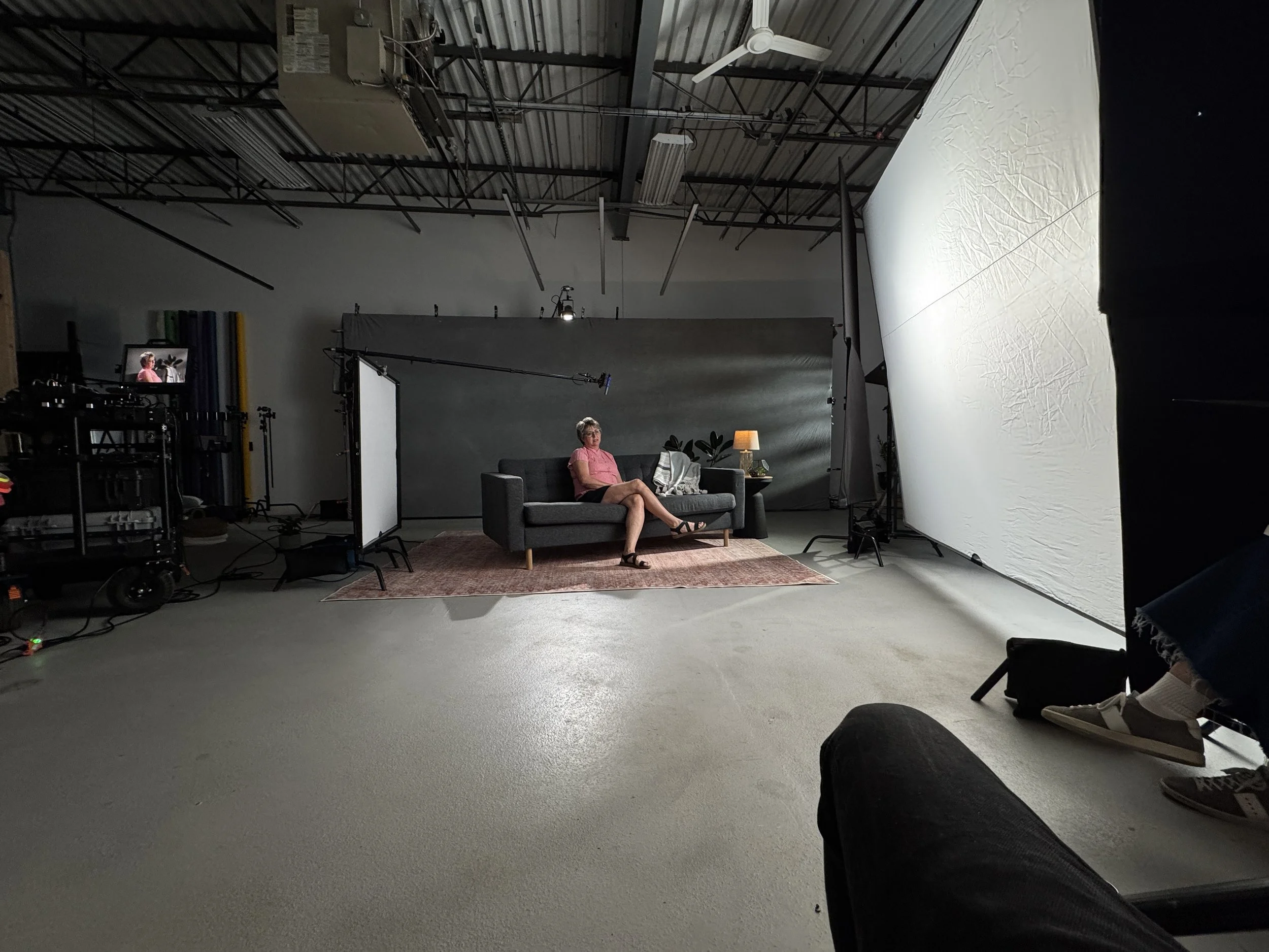 Woman sitting on a dark gray couch in a photography studio set, with a gray backdrop behind her. There are studio lights, a lampshade, plants, and various equipment around the set.