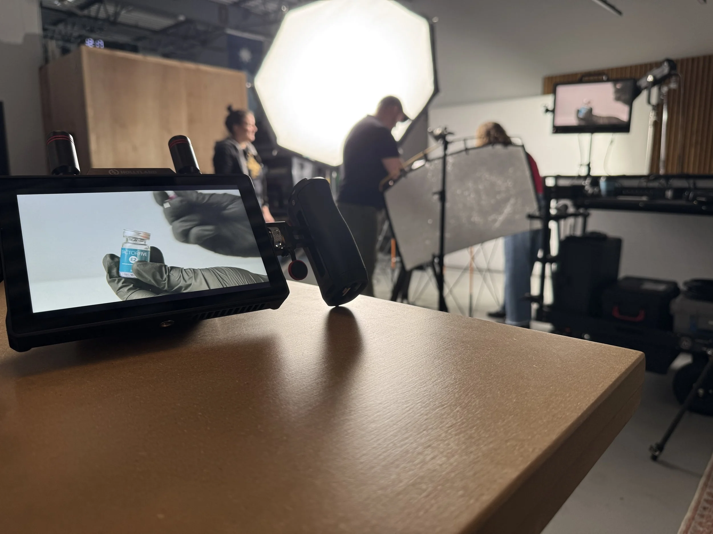 Close-up of a video monitor displaying a person holding a small bottle of medication, with a behind-the-scenes view of a behind-the-scenes of a video production studio in the background, including professionals, equipment, and lighting setup.