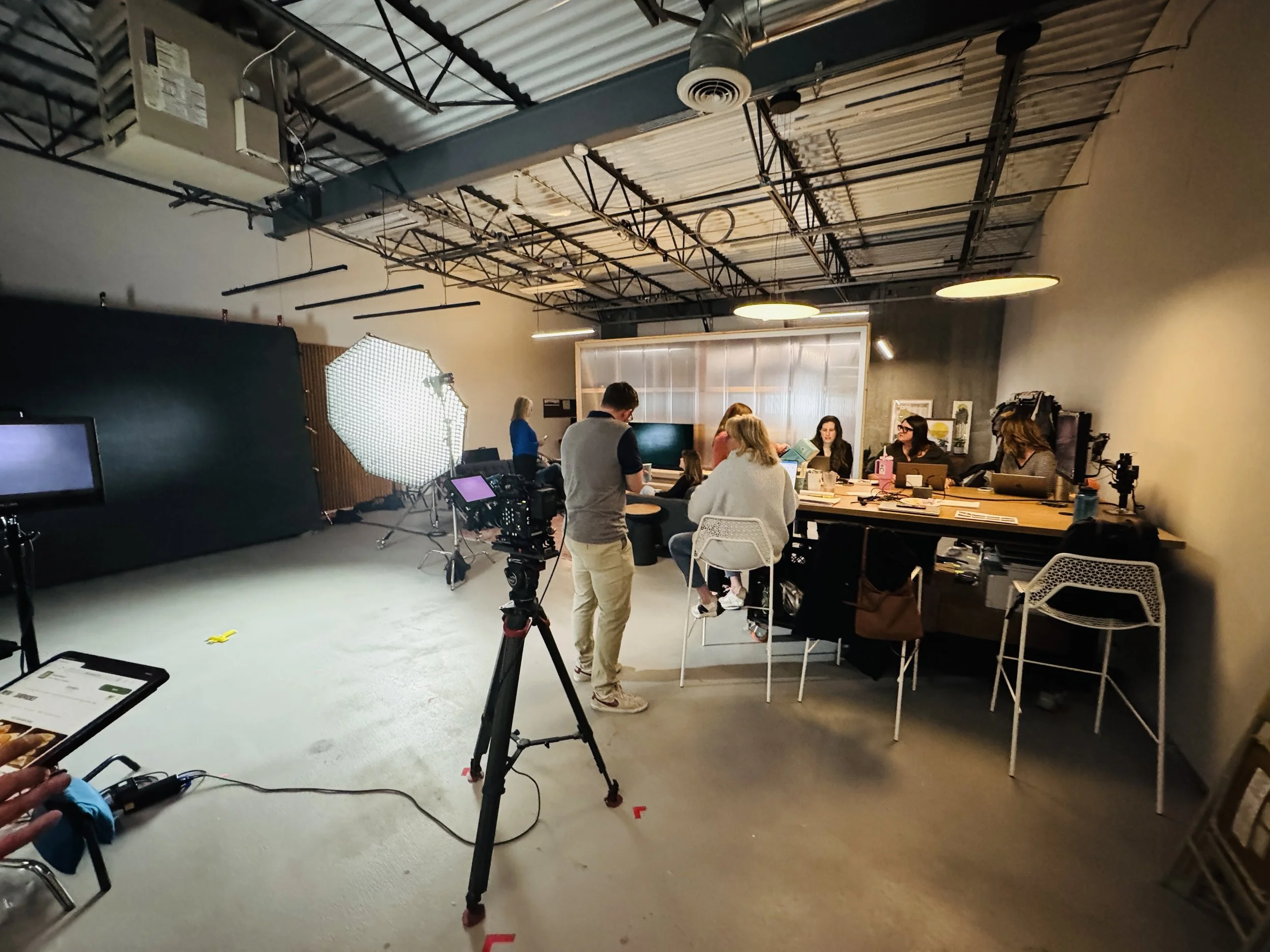 A behind-the-scenes view of a video production or shoot seen in a studio. Several people are gathered around a table with cameras, lights, and filming equipment capturing a scene or interview. The studio has a high ceiling with exposed pipes and a large softbox light on a stand.