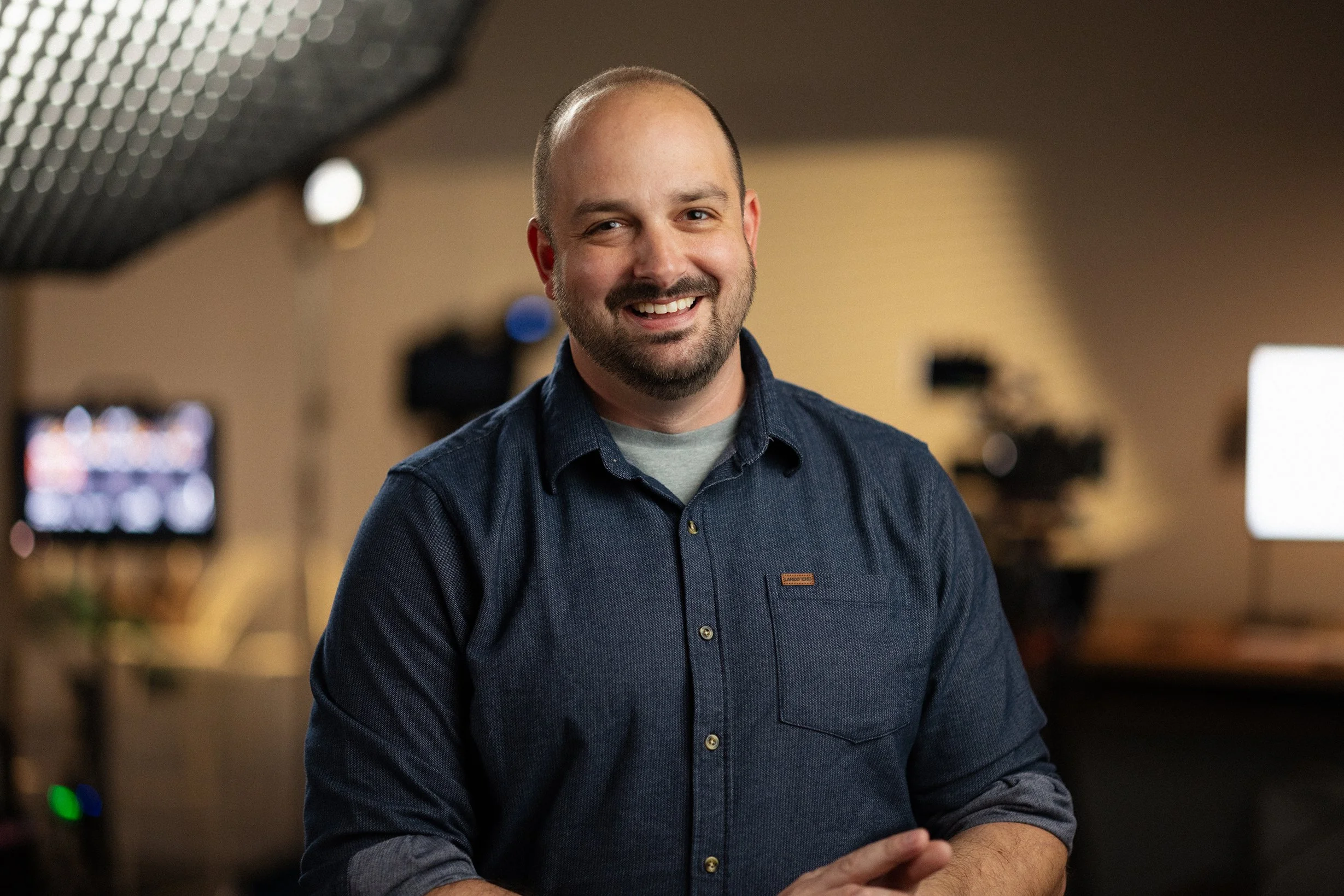 A smiling man with a beard and a denim shirt standing in a professional recording or video studio.