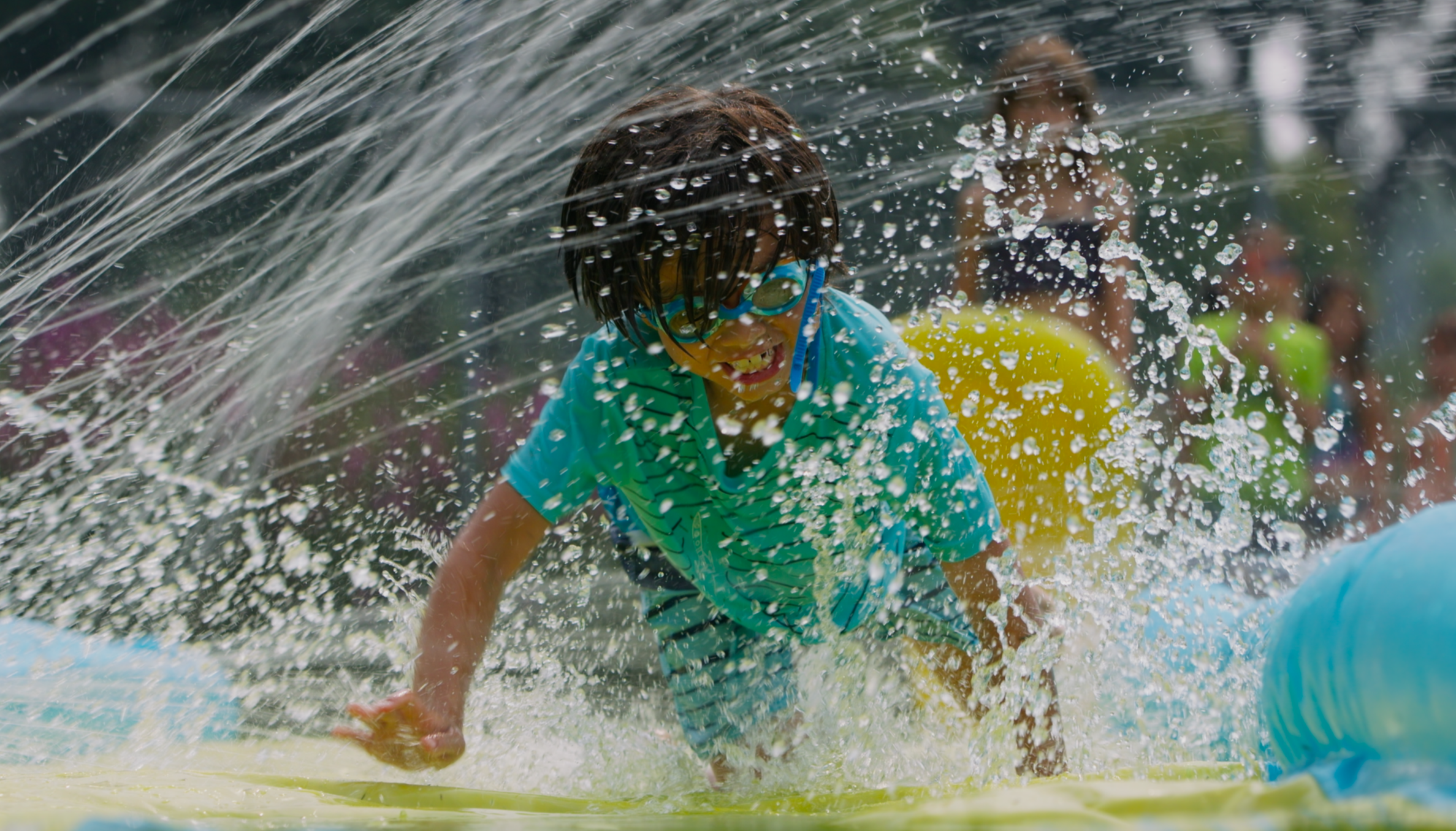 Child wearing goggles and a striped green shirt sliding on a yellow water slide with splashing water, other children in the background.