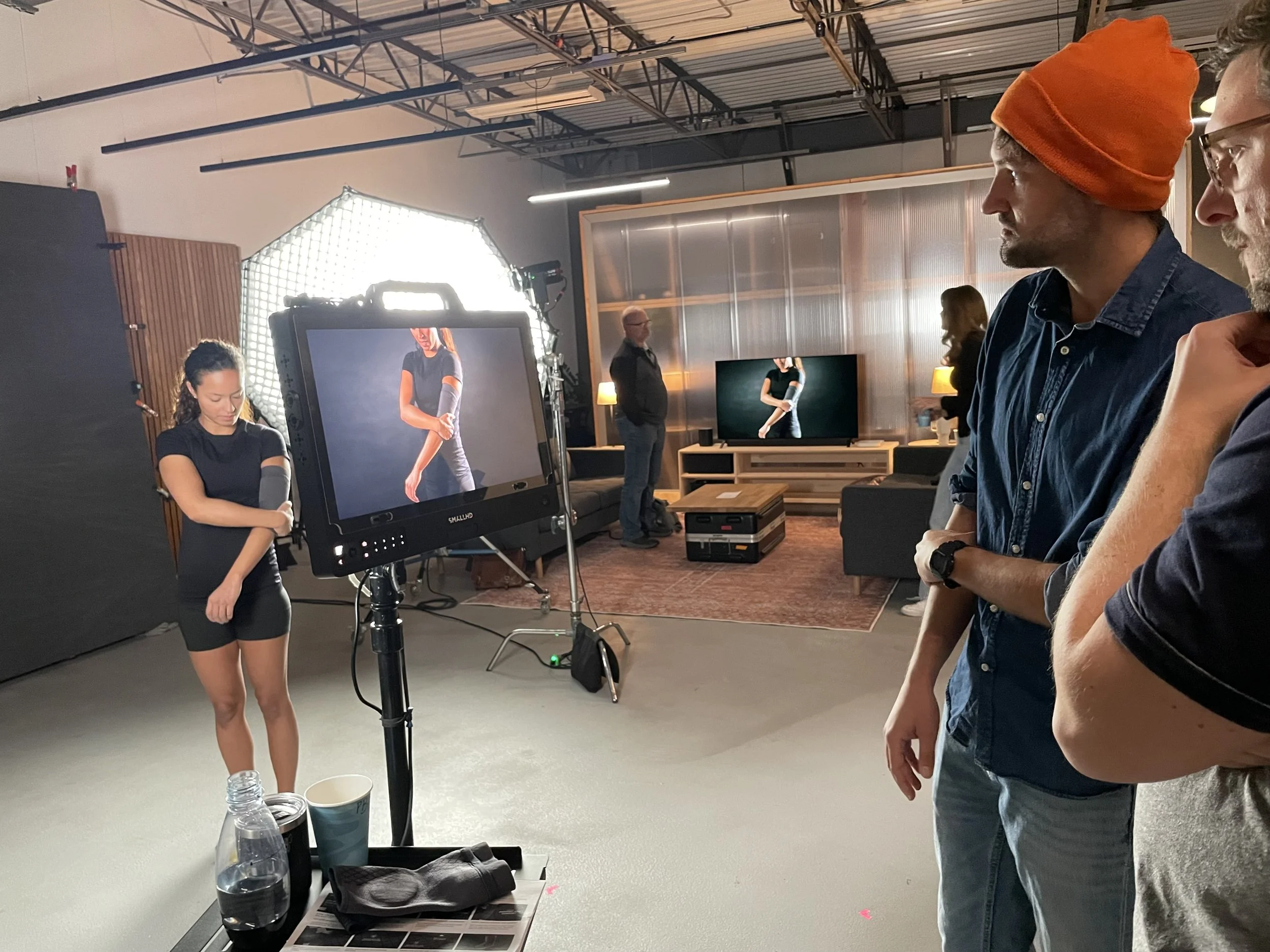 A behind-the-scenes look at a video shoot or photoshoot, featuring a woman in a black outfit with her arms crossed, a monitor displaying a close-up of her arm, crew members watching, and professional lighting equipment in a studio setting.