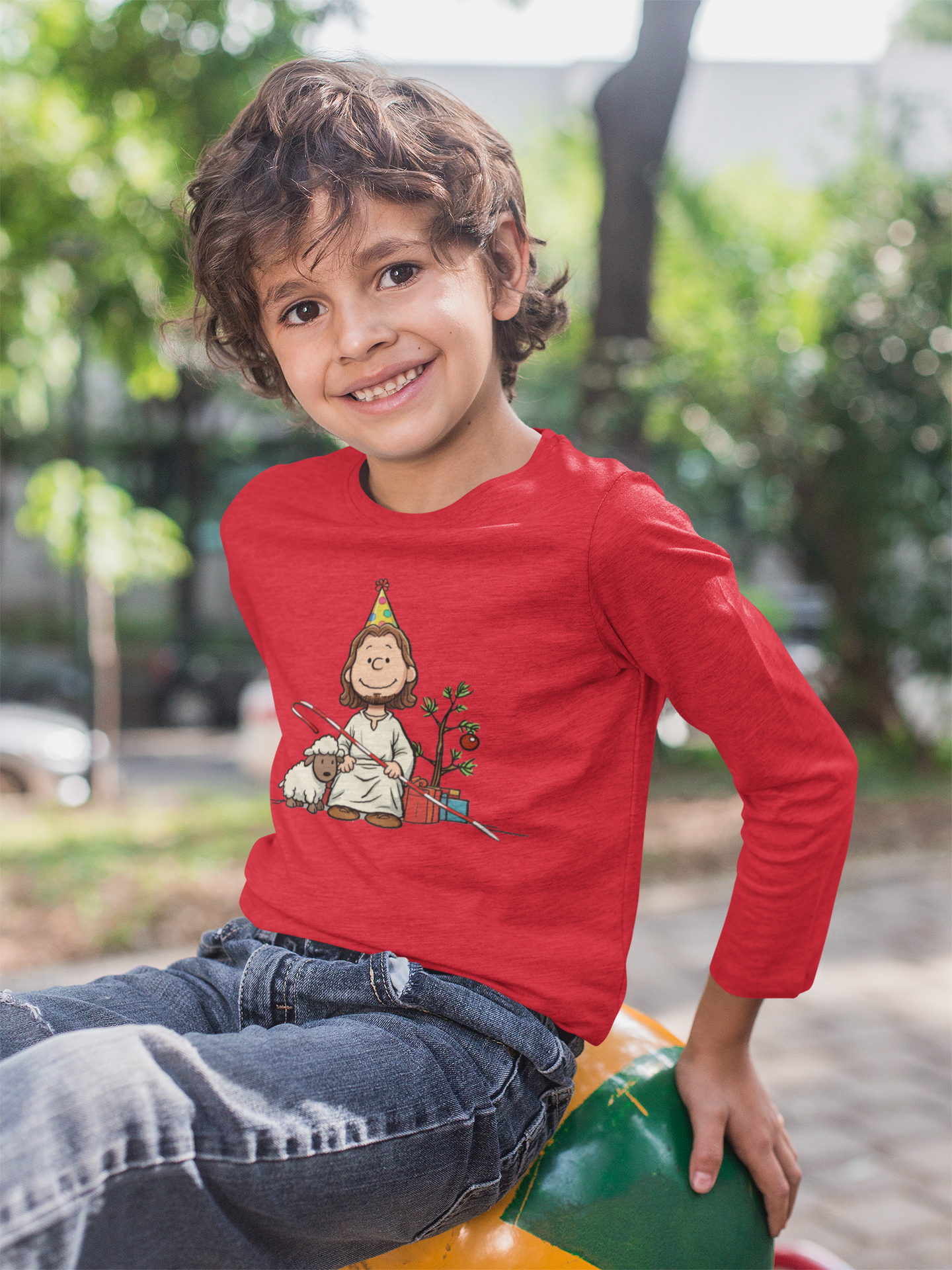 smiling boy at park wearing red toddler long sleeve tee with image of Jesus drawn in Peanuts style with birthday hat and Christmas decor