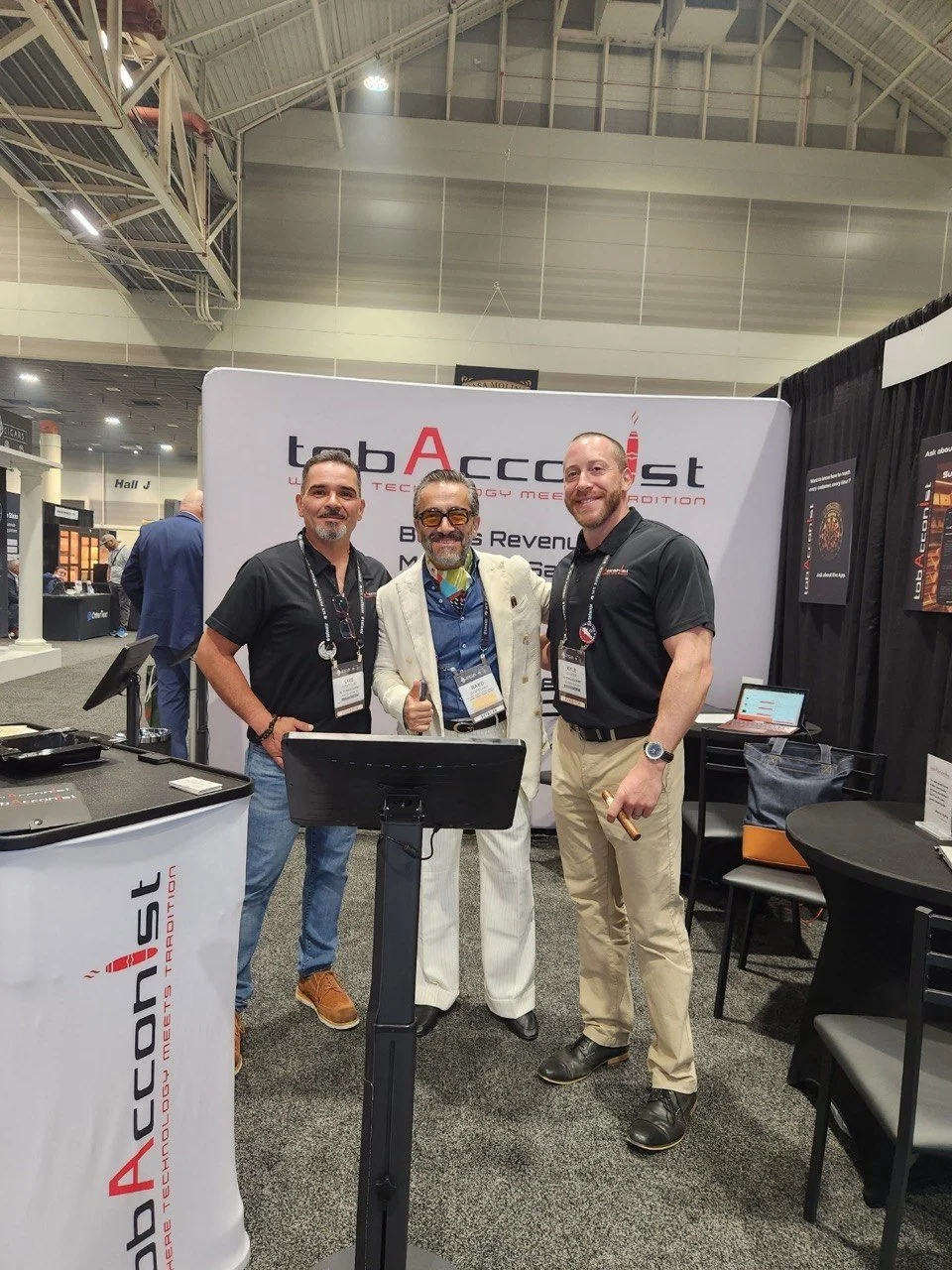 Three men standing at a trade show booth with a backdrop that reads "tabAccent." They are smiling and facing the camera, with the middle man giving a thumbs-up. The booth displays various tech products, and there are tables and chairs nearby in the convention center.