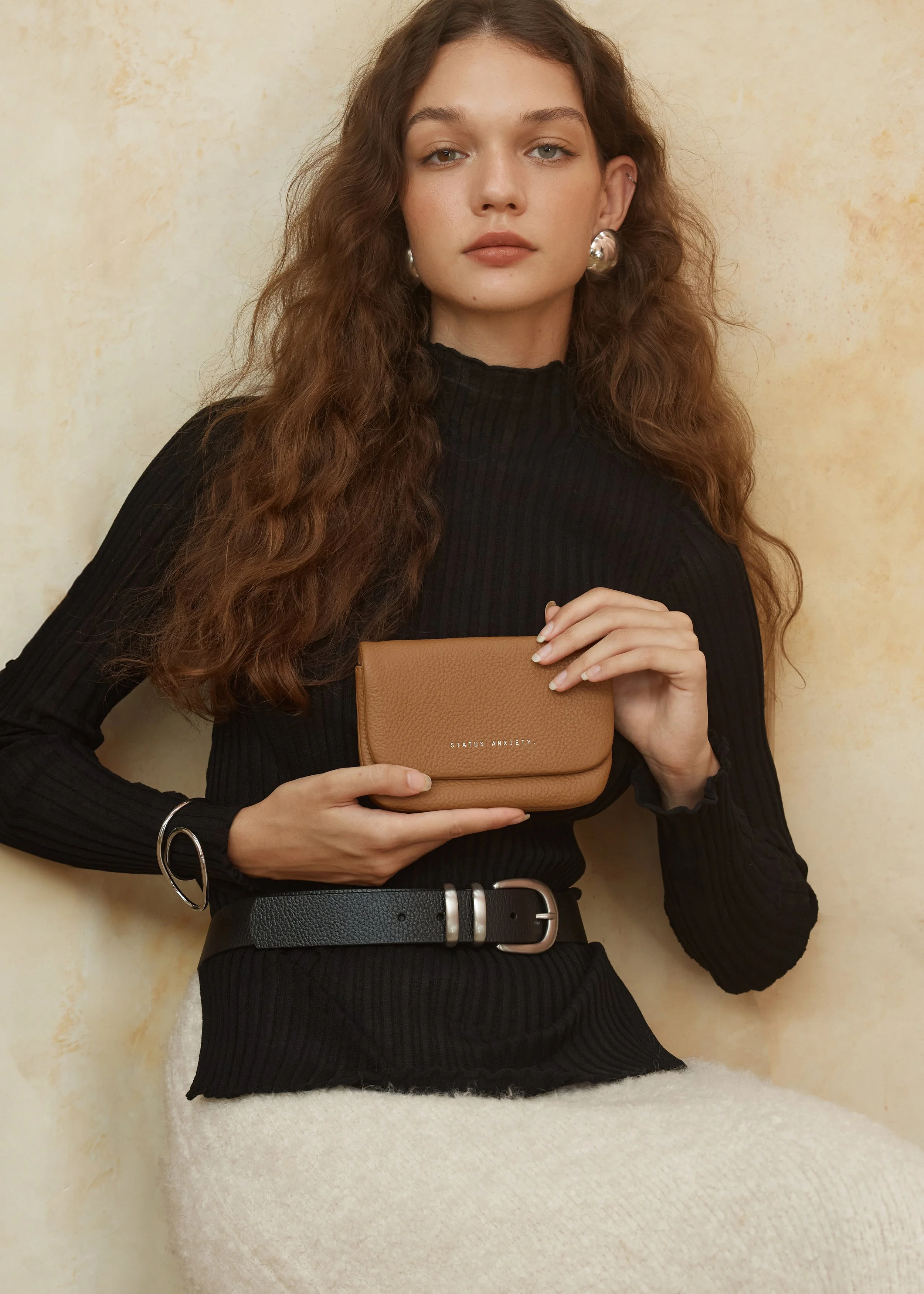 a woman holding a tan leather pouch by "STATUS ANXIETY." She is dressed in a black ribbed top, accessorized with a black leather belt with silver buckles and a silver bracelet