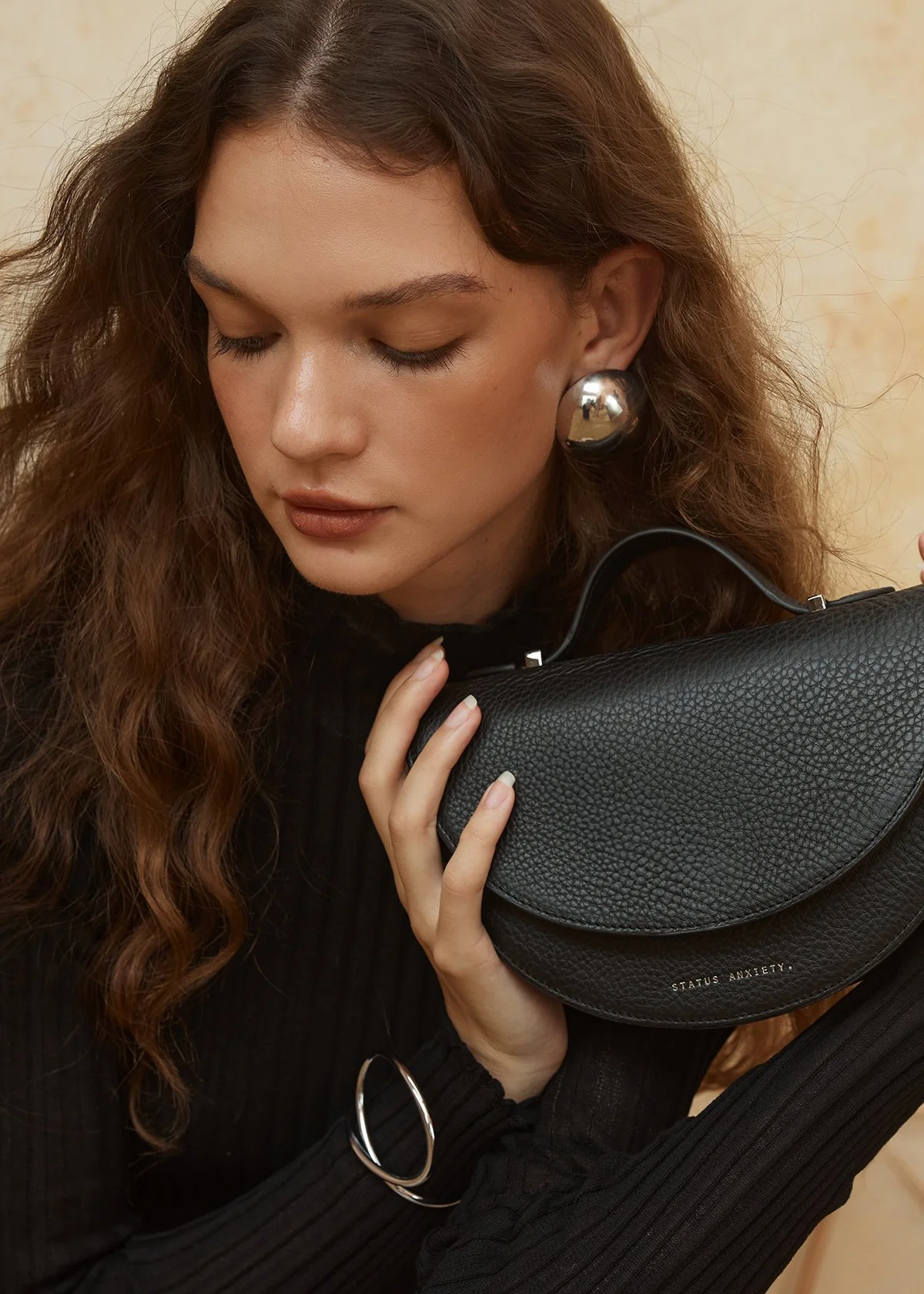 a woman holding a textured black leather bag, branded with "STATUS ANXIETY." She is styled with silver jewelry, including bold spherical earrings and a twisted bracelet