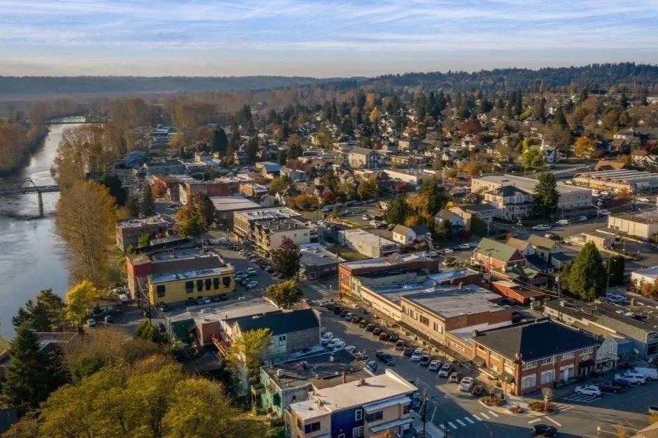 Aerial view of a small town with a river on the left side, trees with fall colors, and a mix of commercial and residential buildings under a partly cloudy sky.