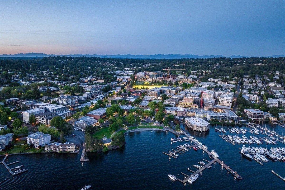 Aerial view of a city waterfront at dusk, showing boats in a marina, a small park, and a dense urban area with buildings and streets extending into the distance.