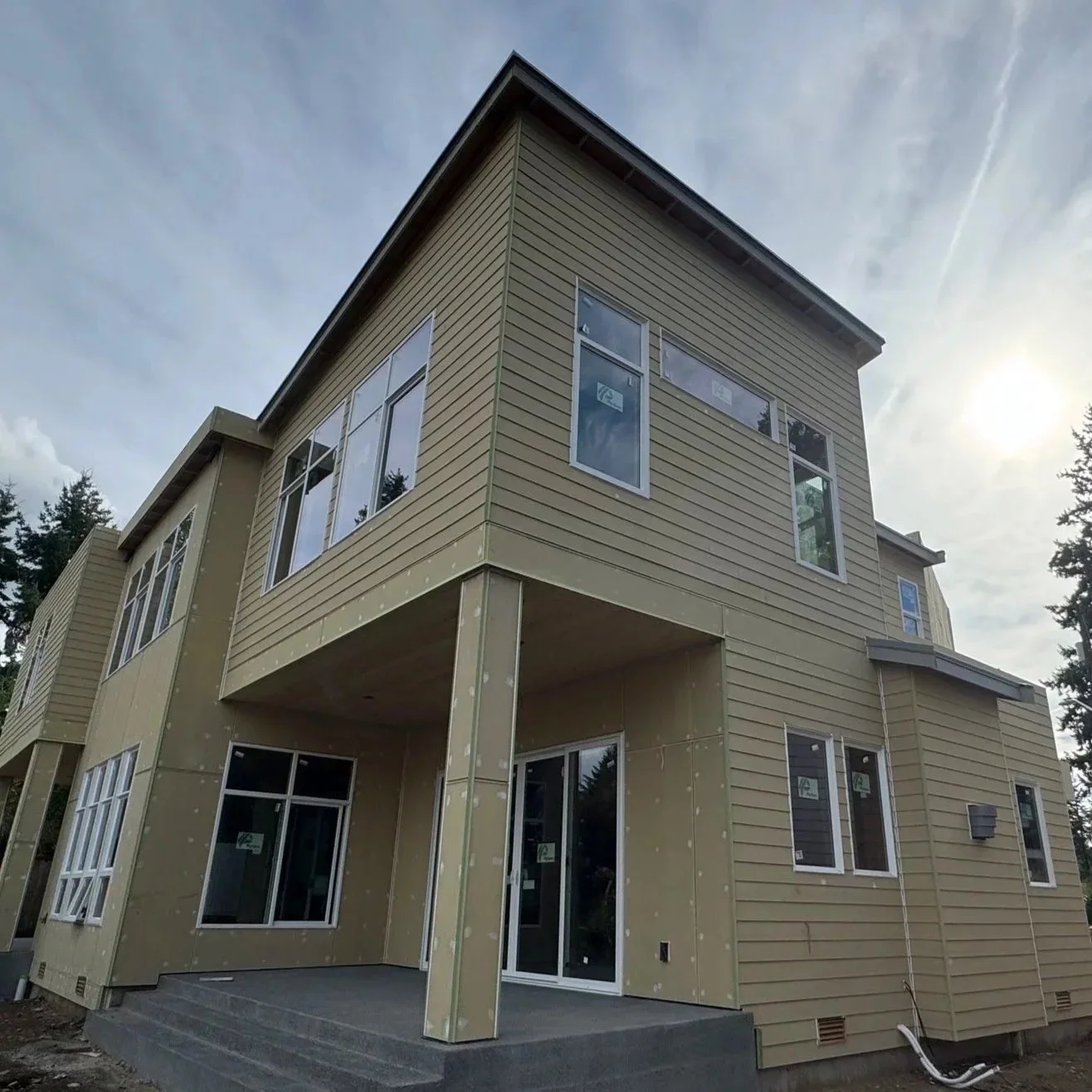 A multi-story house under construction with beige siding, large windows, and a small covered patio area, set against a cloudy sky with the sun partially visible.