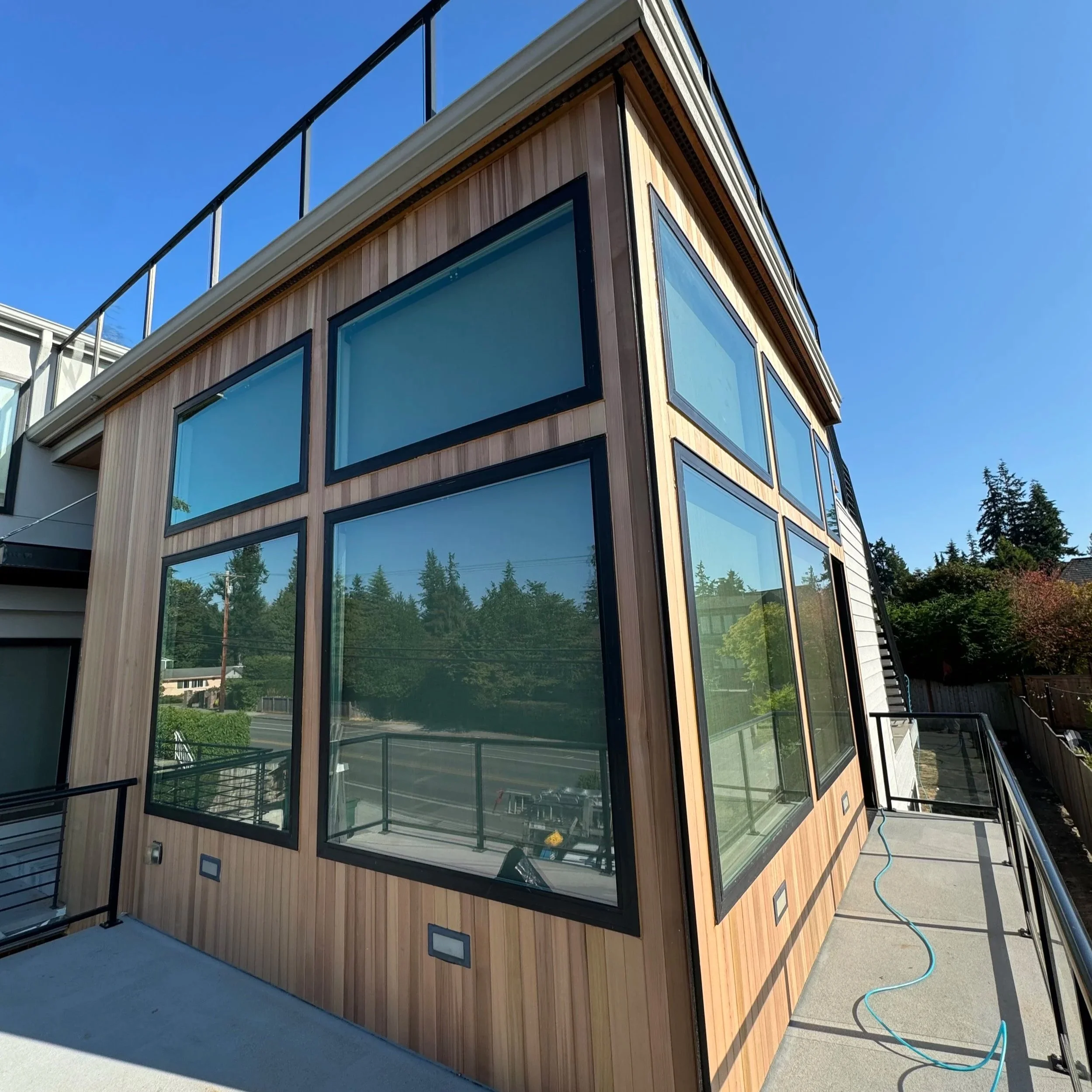 A modern multi-story building with extensive glass windows and wooden paneling exterior. The building is set against a clear blue sky with some trees visible in the background. There is a railing along a nearby balcony and a garden hose lying on the ground.