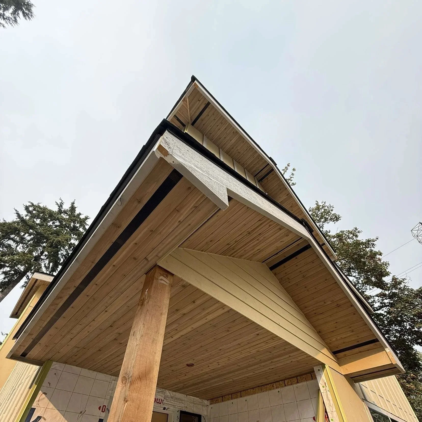 Looking up at an under-construction wooden building, showcasing the roof's underside, support beams, and partially built walls with trees in the background.