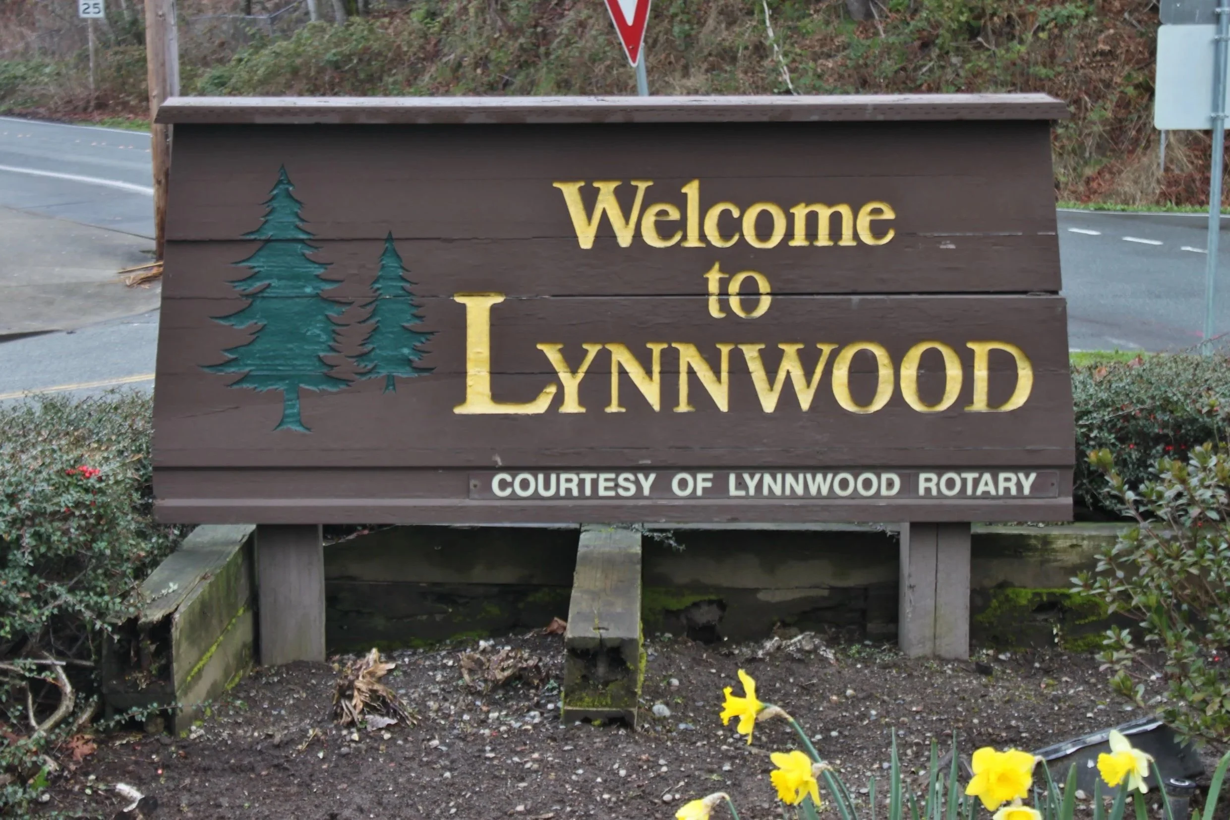 A wooden welcome sign for Lynndood with pine tree illustrations and a courtesy of Lynndood Rotary note, surrounded by flowers and plants, on a roadside.