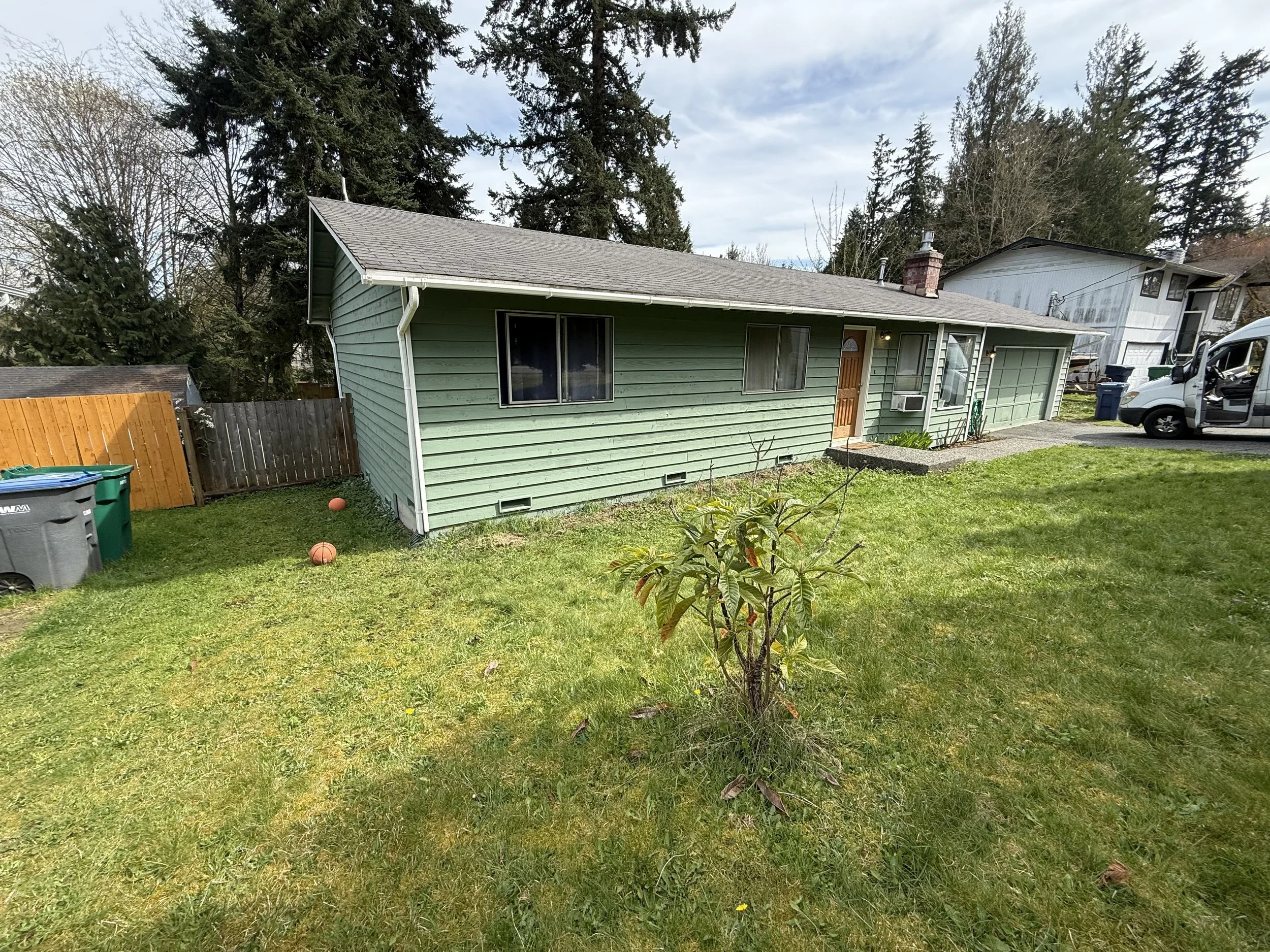 A backyard scene with green grass, a small tree, a wooden fence, and a house with green siding. There are two basketballs on the ground, garbage and recycling bins on the left, and a white van parked on the driveway. Tall trees and other houses are visible in the background under a partly cloudy sky.