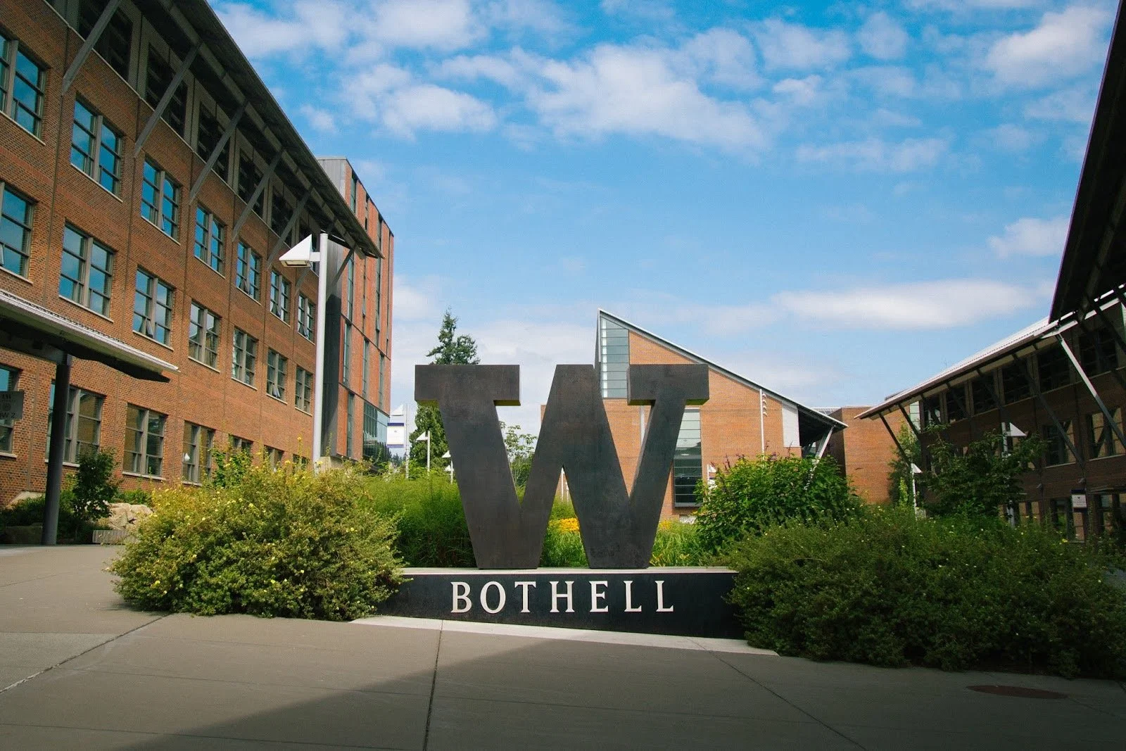 Photo of a university campus with a large metal letter 'W' and the word 'BOTHELL' below it, surrounded by modern brick and glass buildings, greenery, and a blue sky with some clouds.